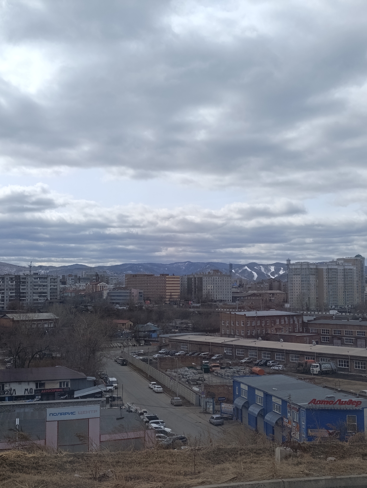 The image depicts a cityscape under an overcast sky. A mountain range is visible in the background, with some snow cover on the peaks. The foreground is dominated by urban structures, including apartment buildings, commercial buildings, and roads lined with parked cars. Signage in Russian script is visible on the commercial buildings. Specific businesses include "Автолидер" and "Поларис Центр". The ground in the immediate foreground appears to be an overgrown embankment or hillside. The setting is Krasnoyarsk, Russia.