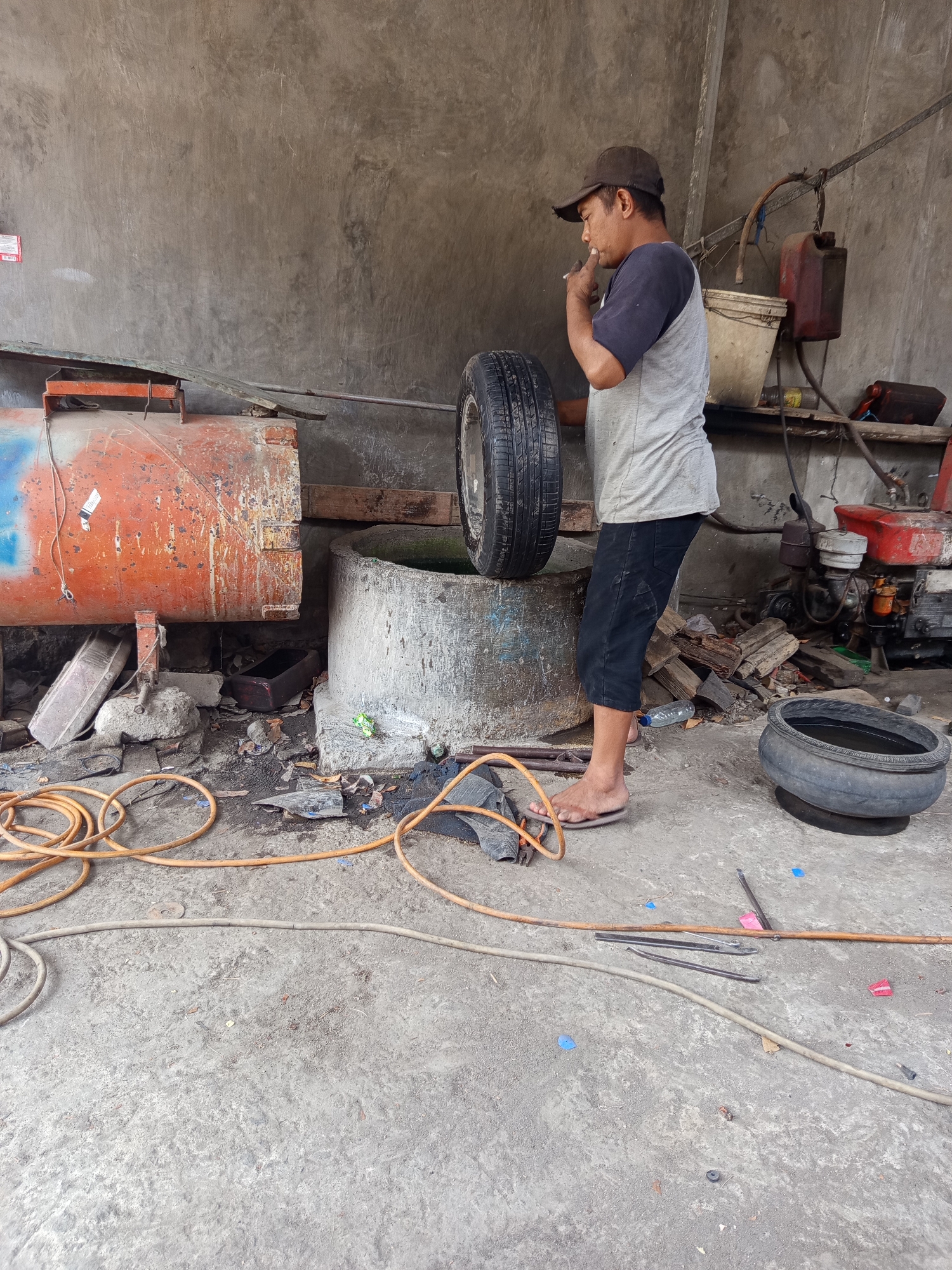 A man in a baseball cap and a t-shirt is standing in what appears to be a workshop or garage area in Mrican, Indonesia. He is holding a tire and smoking a cigarette. The background shows a concrete wall, a large orange cylindrical object, and various tools and equipment, including hoses, a small engine, and buckets. There is also a concrete well or basin filled with water, and a discarded tire. The ground is concrete and littered with debris. The overall impression is of a working, albeit somewhat cluttered, outdoor or semi-outdoor commercial space.