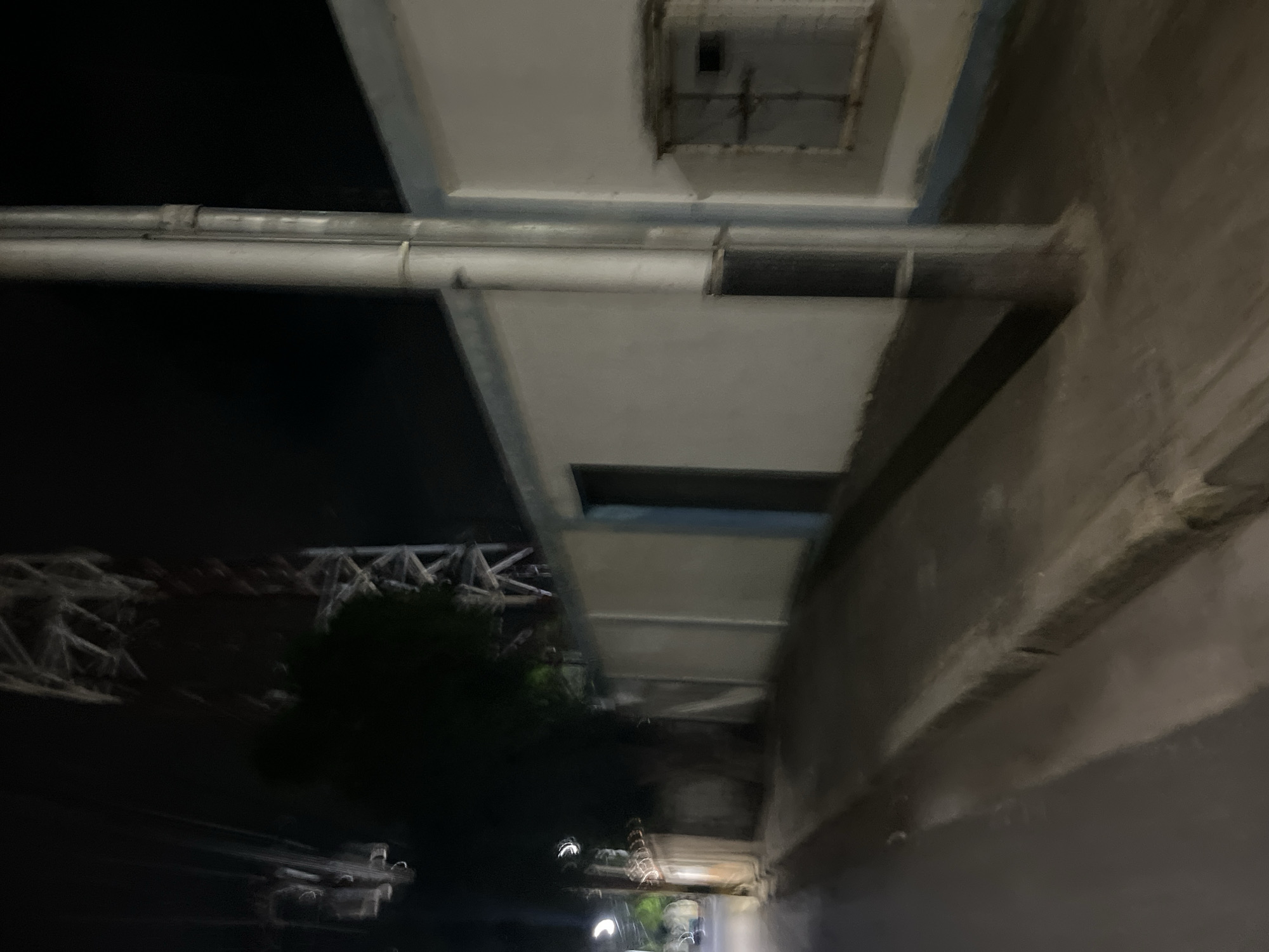 The image depicts a street scene at night in Santa Ana, Venezuela. A tall, lattice-like structure, possibly a radio tower, is visible in the background, partially obscured by darkness and a tree. In the foreground, a white wall runs along the right side of the frame, with a dark, recessed doorway and a small, metal-grated window. A large, vertical pipe runs alongside the wall. A sidewalk runs parallel to the wall, and a paved street is to the left, with some streetlights visible. The overall impression is one of a quiet, possibly industrial or residential area at night.