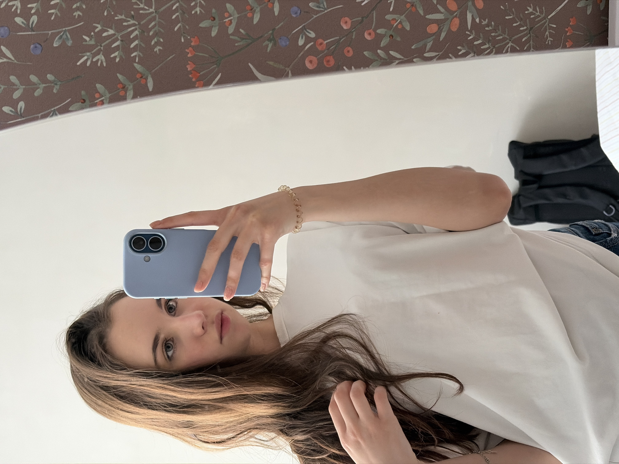 A young woman is taking a selfie in front of a mirror. She is holding a light blue smartphone in her left hand, the camera pointed towards her face. She has long brown hair and is wearing a white t-shirt. A gold bracelet is visible on her left wrist. Behind her, a dark floral pattern decorates the top of the mirror frame, and a dark garment and blue jeans are visible in the reflection. There is no indication that the picture was taken in Izhevsk, Russia.