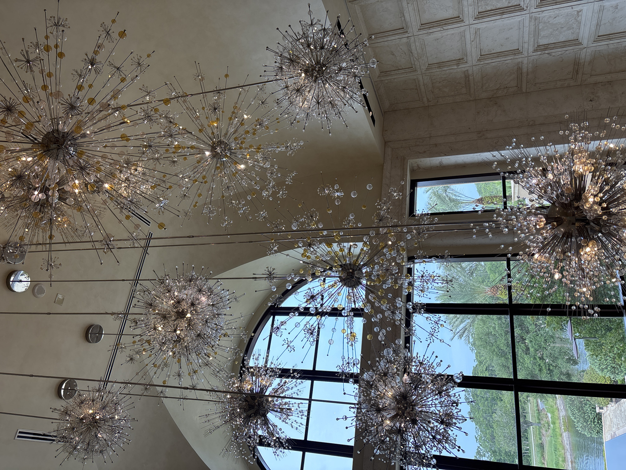 The image captures a view looking upwards inside what appears to be a high-ceilinged room. Several elaborate, dandelion-shaped chandeliers are suspended from the ceiling. Sunlight filters through a large, multi-paned window, revealing a view of trees and foliage outside. The ceiling itself has a textured or patterned finish. The location appears to be in Bay Lake, United States.