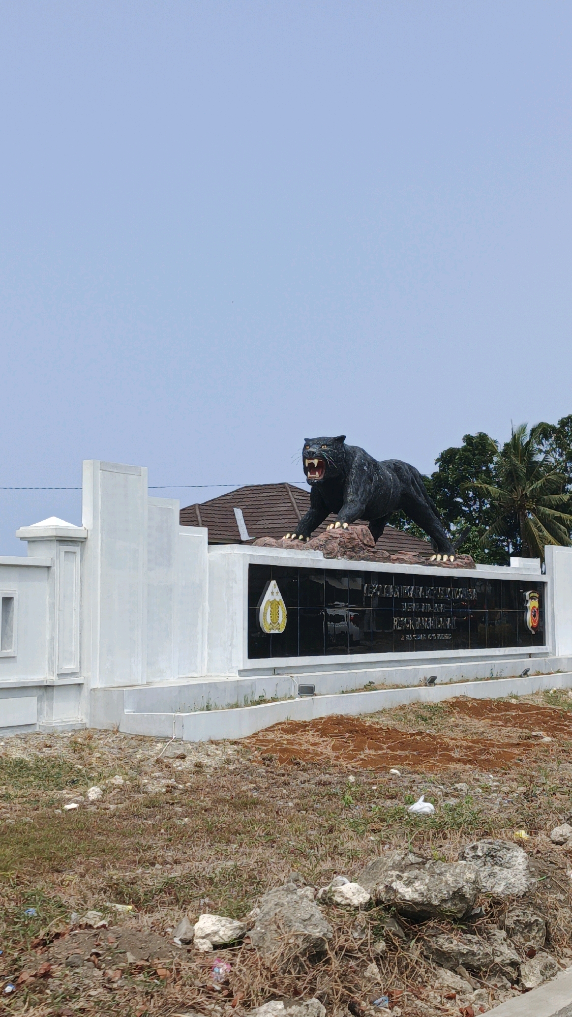 A black panther statue stands aggressively on a rocky base, its mouth open in a snarl, with its front paws planted firmly. The statue is positioned atop a polished black wall with several emblems. To the left of the panther statue, there is a white concrete structure with decorative pillars and a sloped roof. Behind these structures, a building with a brown tiled roof and lush green trees, including a palm tree, are visible. The foreground consists of dry grass and scattered rocks, suggesting an undeveloped or arid environment. The sky is clear and light blue. The scene appears to be located in Desa Wonoharjo, Indonesia, as indicated by the text on the wall, which includes the words "POLRES KEBUMEN" and what appears to be a regional map.
