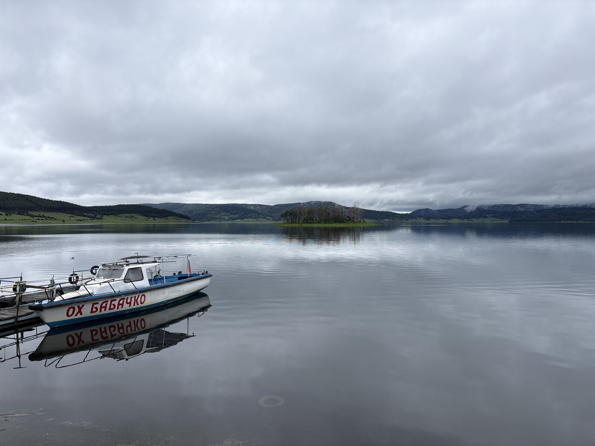 The image depicts a tranquil scene at Lake Batak in the city of Цигов чарк, Bulgaria. A white motorboat, bearing Cyrillic script in red ("ОХ БАБАЧКО"), is docked on the left side of the frame. The boat's reflection is clearly visible in the still water. The lake stretches out towards a backdrop of rolling, tree-covered hills and mountains. A small island with trees is visible in the middle of the lake. The sky is overcast with dense, gray clouds. The overall impression is one of calmness and solitude.
