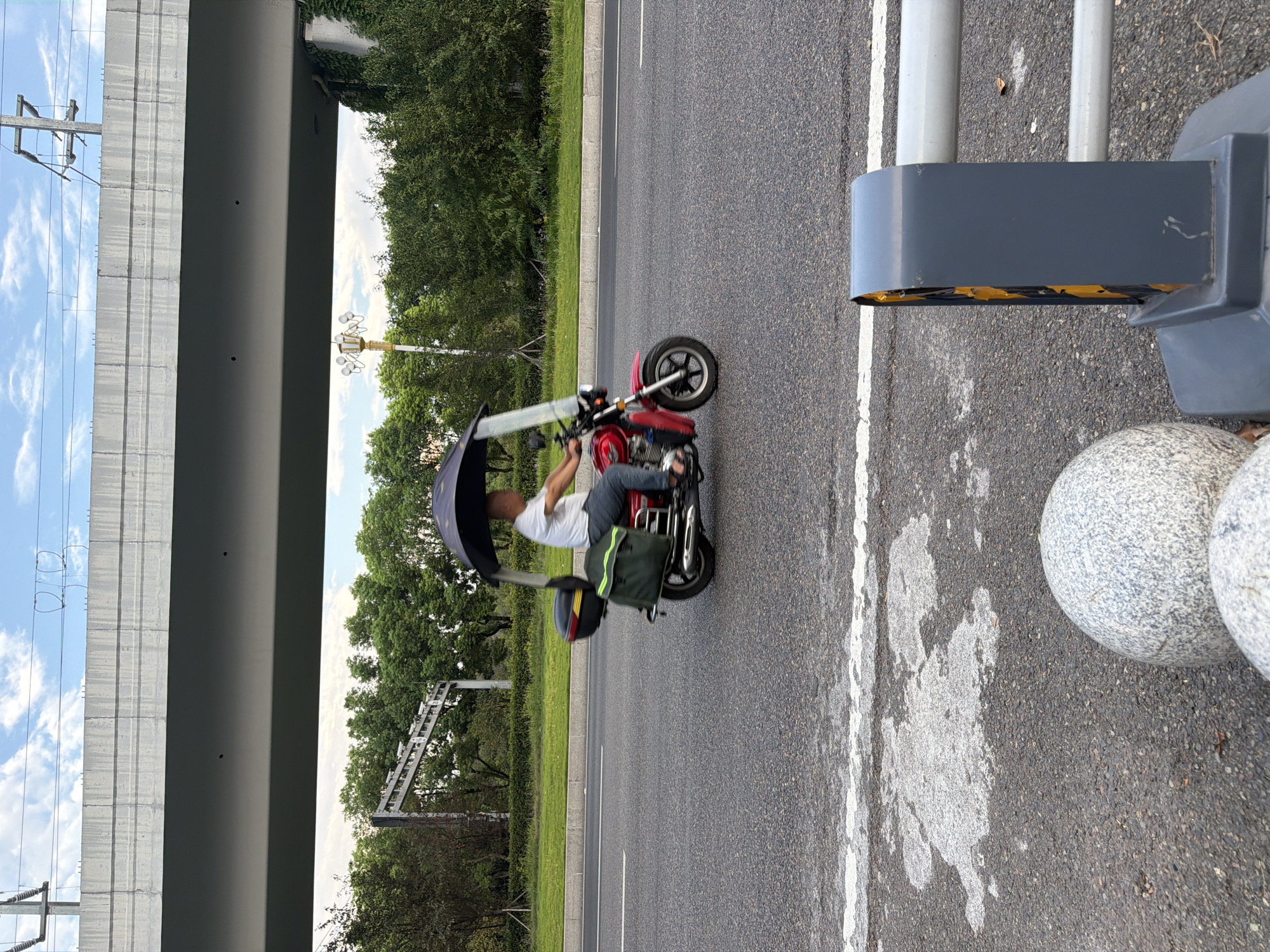 A middle-aged man is riding a red motorcycle with a black canopy and a green saddlebag on his right side, on an asphalt road under an overpass. The man is wearing a white t-shirt and blue jeans. He is looking ahead and holding the handlebars. The motorcycle has a black top box. In the background, there are trees and a street lamp. The overpass is a concrete structure with overhead power lines. The road has a white line marking the lane. In the foreground, there is a gray barrier with yellow accents and two round gray stones. The sky is partly cloudy. This scene is captured in the city of Jiangjin, China.