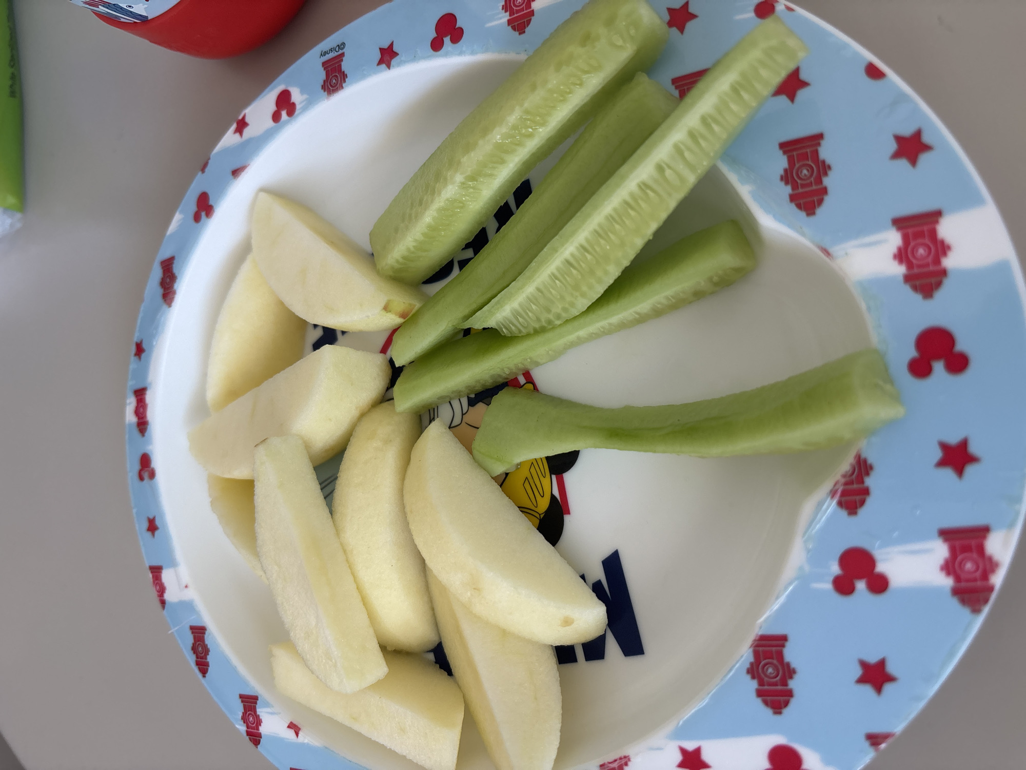 A plate of cut apples and cucumbers is shown on a table. The plate has a blue border with red images of Mickey Mouse ears, stars, and fire hydrants. The apples are sliced into wedges and are light yellow. The cucumbers are cut into sticks and are green. The background is a light-colored surface. There are no people or animals visible in the image. The scene is not in Dubai, United Arab Emirates.
