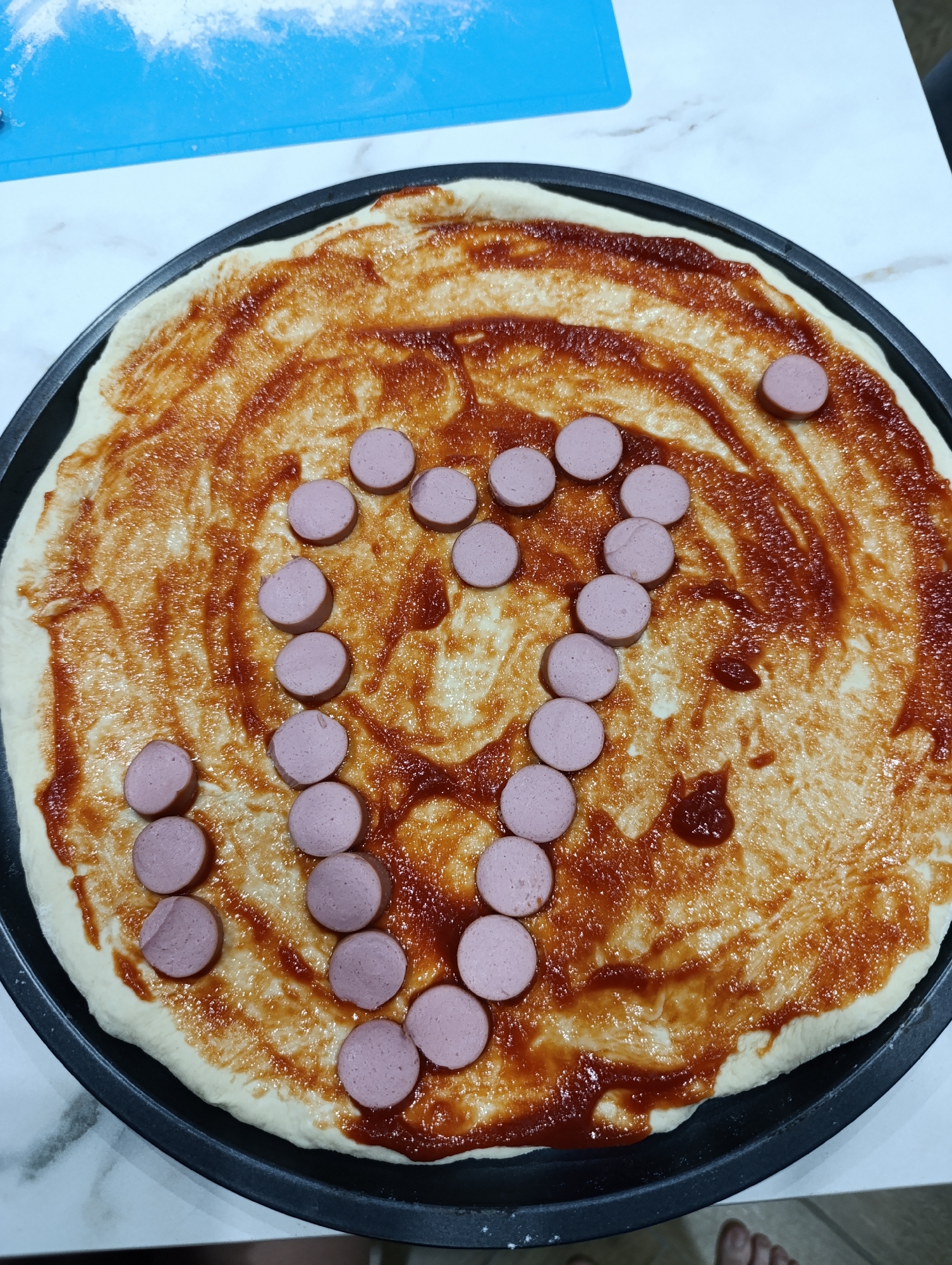 The image shows a homemade pizza on a black pan, likely in a residential setting. The pizza dough is spread with what appears to be tomato sauce. Sliced sausage pieces are arranged to form a question mark in the center of the pizza. A blue cutting board with flour residue sits nearby on a white surface. The photo was likely taken in a kitchen, based on the countertop surface. While not explicitly verifiable, given the image origin, it is plausible to infer the location as Chaikovsky, Russia.