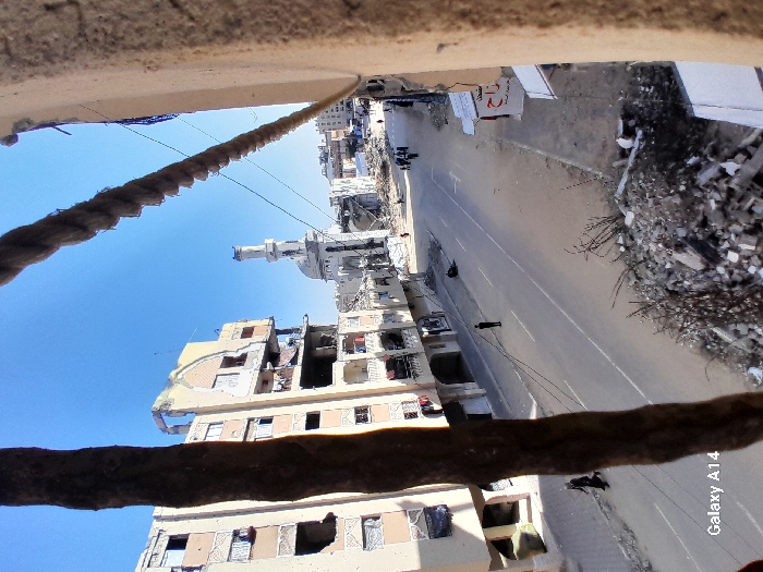 The image captures a street scene in Khan Yunis, Palestinian Territory, under a clear blue sky, revealing extensive urban destruction. In the immediate foreground, a dark vertical object (likely a tree trunk or beam) partially obscures the view on the left, while a thick rope or cable spans the upper right. Piles of rubble, concrete fragments, and twisted debris litter the bottom edge and extend along the sidewalks.
A paved street, marked with two white lines, runs through the center, largely clear of major debris. On the left side of the street, a multi-story building (approximately 6-7 floors visible) shows severe damage, with large sections of its facade missing, exposing internal rooms, and numerous blown-out windows. One individual, dressed in dark clothing, walks on the sidewalk next to this damaged building. Further down the street, another person in dark attire stands alone in the middle of the road.
In the midground, a mosque with a prominent, slender minaret stands heavily damaged; its dome and surrounding structures exhibit significant destruction, with large sections reduced to rubble. A group of approximately five to seven individuals walks together on the right side of the street, nearing the mosque. Further down the street on the right, a dark object lies on the ground.
Beyond the mosque, the background reveals more devastated buildings and widespread rubble, indicating further damage to the urban landscape. Several utility wires or cables are strung across the scene overhead. A "Galaxy A14" watermark is visible in the bottom-left corner.