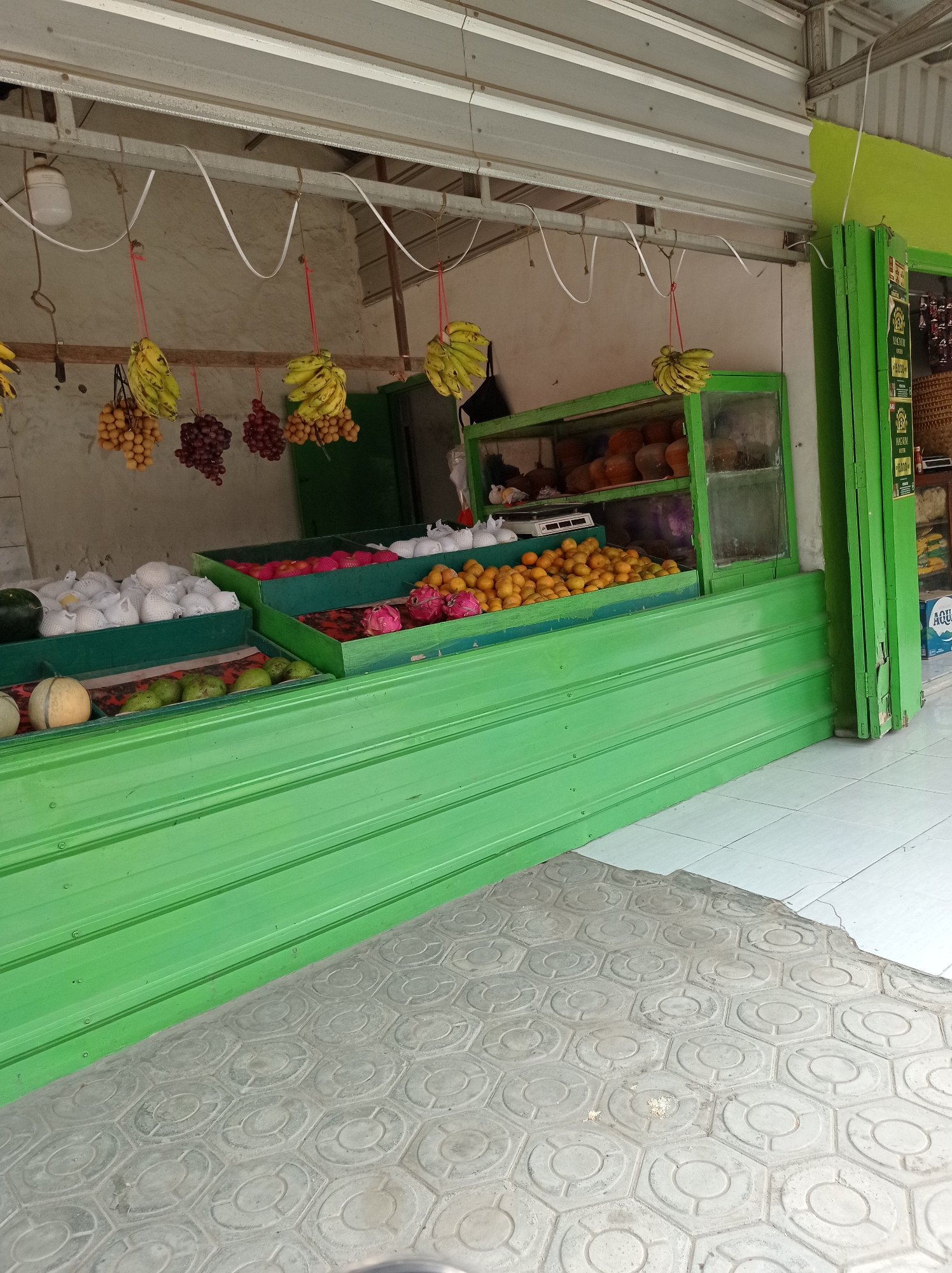 This is a medium shot, eye-level, outdoor photograph of a fruit stall located in the city of Kramat, Indonesia. The stall is brightly painted in green and has a covered roof. Hanging from the roof are bunches of bananas, grapes, and other yellow fruits. In the foreground, a green counter displays various fruits, including what appear to be dragon fruit, oranges, watermelons, melons, and avocados. Behind the counter, there is a glass display case filled with more fruits and some terracotta pots. The ground in front of the stall is paved with hexagonal tiles. The overall scene depicts a typical street-side market setting in Indonesia.