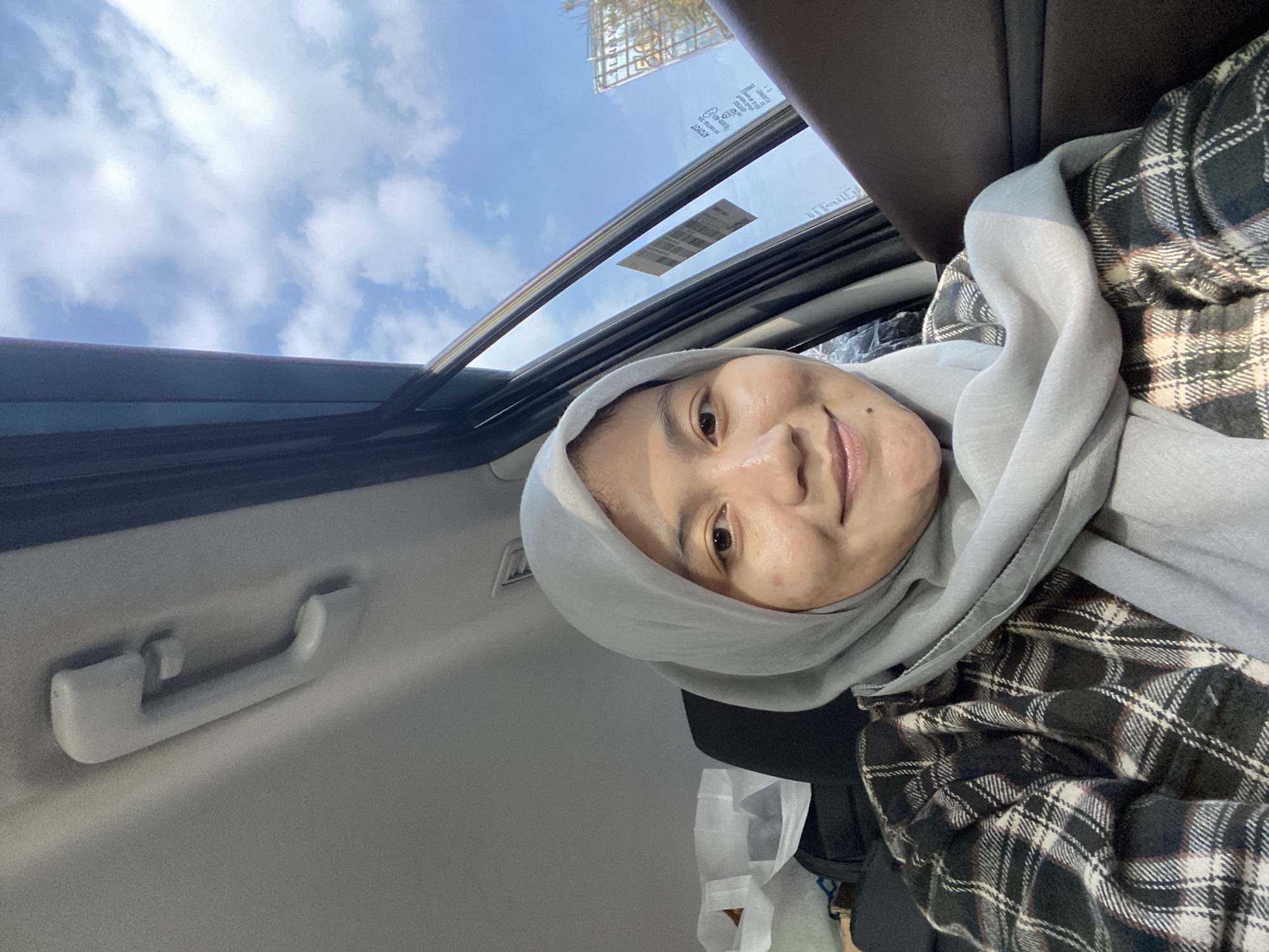 A young woman wearing a light gray hijab and a plaid shirt is seated in the passenger seat of a car. She is looking towards the camera and smiling. Sunlight streams in through the car window, illuminating her face. Outside the window, a blue sky with scattered clouds and a billboard are visible. In the background inside the car, a white shopping bag and dark luggage are present. The image appears to be taken in Mandar Sari, Indonesia, given the presence of typical Indonesian billboards and architectural styles.