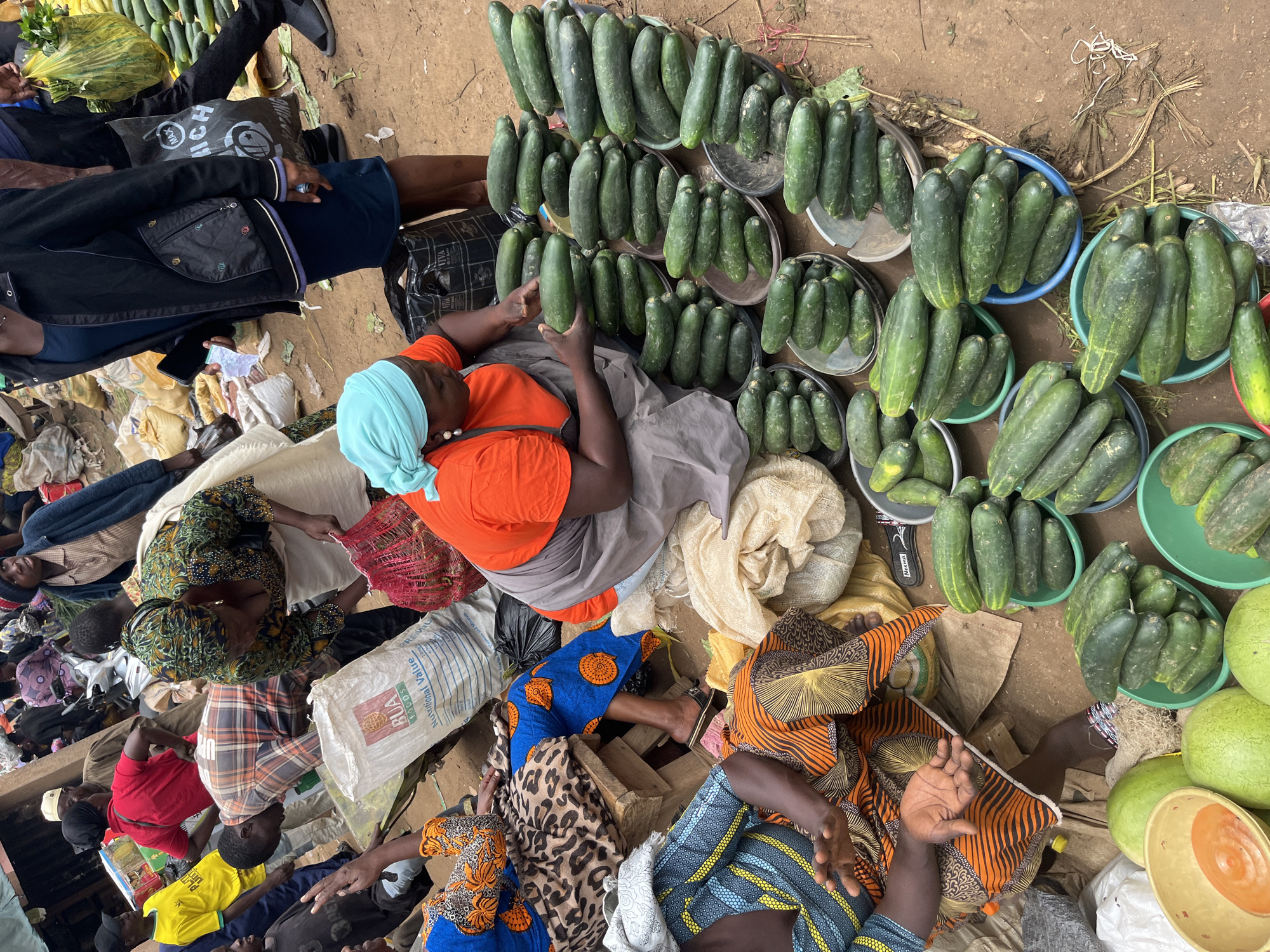 This image depicts a bustling outdoor market scene, likely in Gura Top, Nigeria. The foreground is dominated by an abundance of fresh cucumbers, displayed in various shallow bowls and piled high. Several people, primarily women, are interacting with the produce. One woman in an orange shirt and blue headscarf is carefully examining a cucumber, while others, some in vibrant patterned clothing, are visible in the background. The ground is dirt, typical of an outdoor market. Bags of produce and other market goods are scattered throughout the scene, indicating a lively commercial environment.