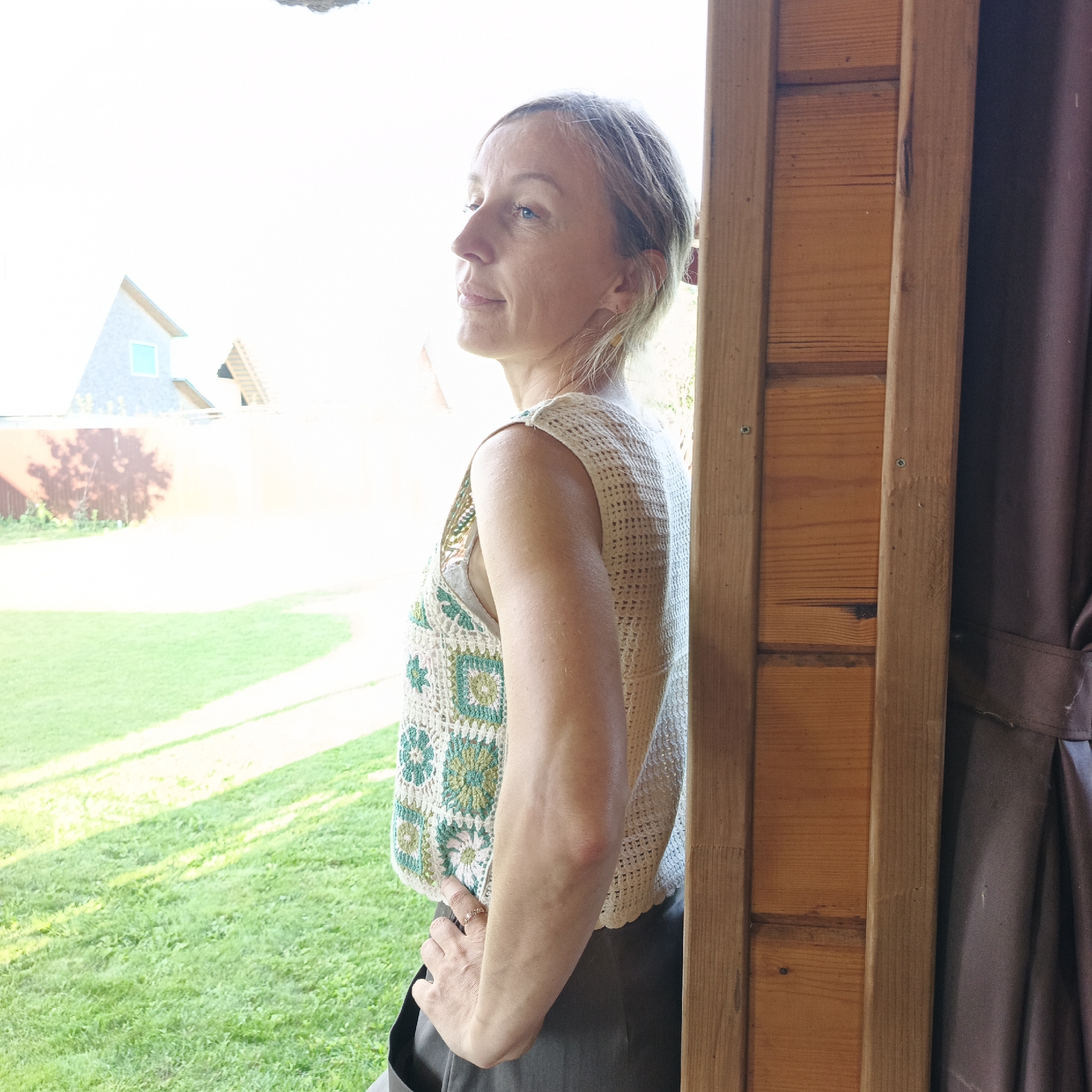 A woman stands in a doorway, looking out at a sunny, grassy yard with a house in the background. The woman is wearing a cream-colored crocheted vest with green floral patterns over a light-colored camisole and gray pants. Her blonde hair is pulled back. Her left hand is on her hip, and she is wearing a ring. The doorway is made of wood. The house in the background is light-colored with a gray roof and a small blue window. The location is the city of Chaikovsky, Russia.