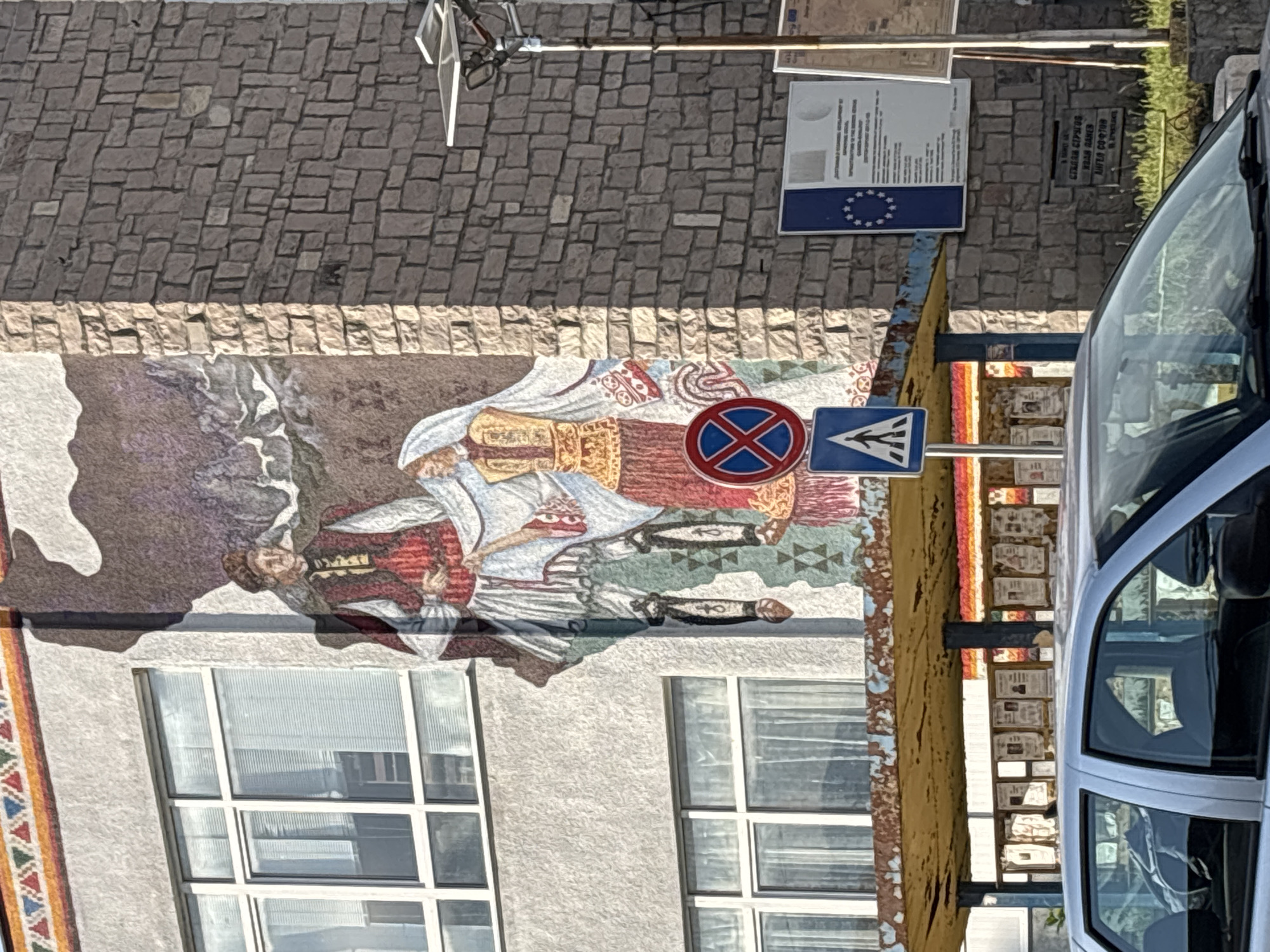 The image shows a building in Garmen, Bulgaria, featuring a large mural. The mural depicts two figures in traditional Bulgarian attire against a mountain backdrop. Adjacent to the mural are street signs: a "no stopping" sign (red circle with a blue "X") and a pedestrian crossing sign (blue triangle with a white figure). Part of a light gray car is visible on the right side of the image, partially obscuring the scene. Above the mural are brick and stone walls. Further up are street signs with information about Garmen.