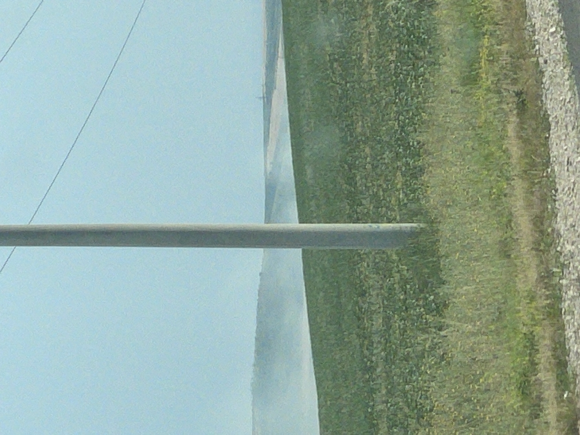 The image is a landscape oriented photograph with a vertical tilt. The scene appears to be outdoors in a rural or semi-rural setting, possibly near Comrat, Moldova. The dominant features are a large, grey cylindrical object running horizontally across the middle of the frame, likely a pipe or beam. To the left of this object is a vast expanse of pale blue sky, with two thin, dark lines that could be wires or power lines stretching across it.
To the right of the pipe, the scene transitions to a dense, green field, suggesting a crop or vegetation. The field occupies the majority of the right side of the image and stretches towards the background. Along the far right edge of the image, there's a strip of grey, gravelly ground, which could be a road or path.
In the middle ground, a thin plume of white mist or smoke is visible, rising from the field and obscuring a portion of the transition between the sky and the field. This plume creates a hazy effect. The lighting suggests it is daytime, likely with clear skies based on the color of the sky. There are no people or animals visible in the frame. No text is discernible in the image. The overall impression is one of an agricultural landscape with some industrial or infrastructural elements present.