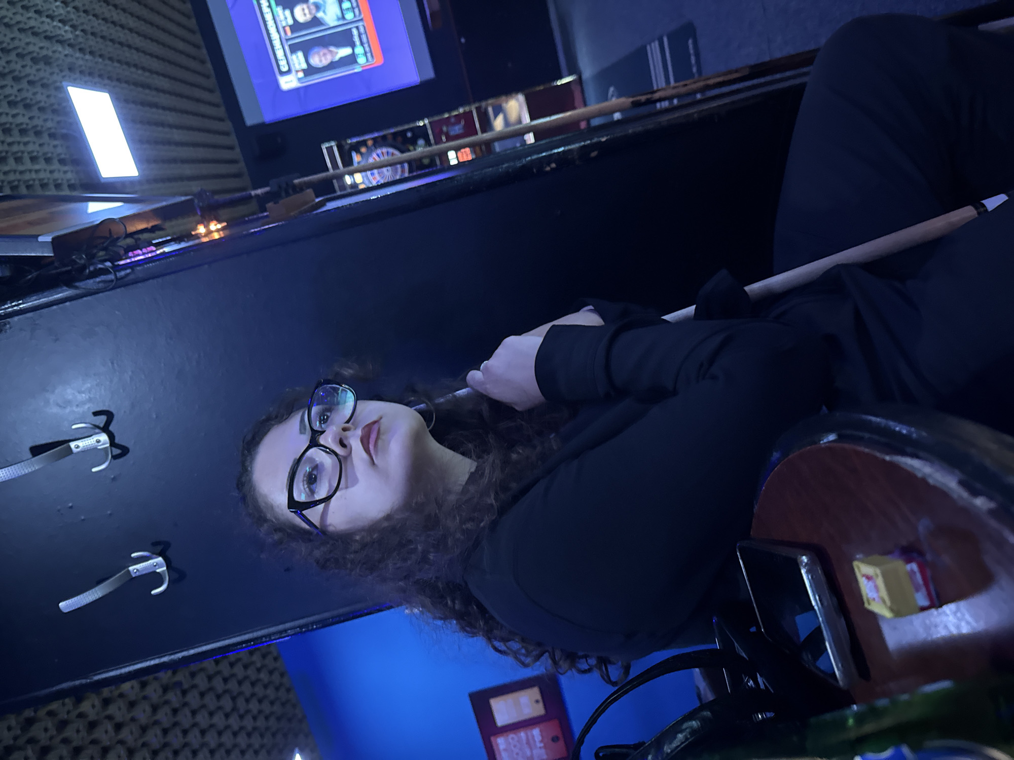 A woman with curly dark hair and glasses is sitting at a wooden table, holding a pool cue. She is wearing a black long-sleeved shirt. On the table in front of her are a smartphone, a small yellow and red candy, and a portion of a green bottle. The background shows a blue wall with what appear to be coat hooks, a dartboard, and a television screen displaying what looks like a game or competition. The overall setting suggests an indoor entertainment venue, possibly a pool hall or bar. The location is Yambol, Bulgaria.