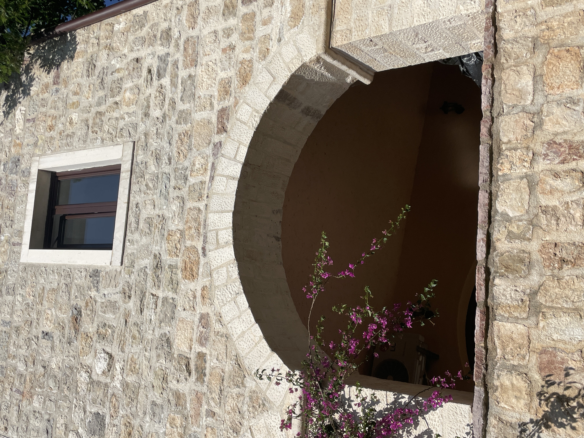 The image shows a stone wall with two window openings. One is a rectangular window with a brown frame. The other is a larger, arched opening. Purple bougainvillea flowers are growing in front of the arched opening. The wall appears to be part of a building in the city of Budva, Montenegro. Inside the archway is a terracotta-colored wall, a dark object wrapped in plastic hanging, a light fixture, and a modern air conditioning unit.