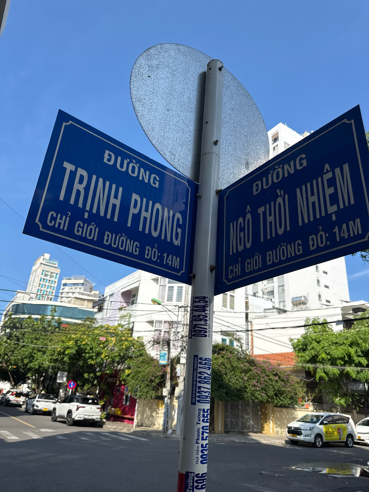 The image shows a street scene in Nha Trang, Vietnam, featuring a blue street signpost with two signs indicating street names. The top sign on the left reads "ĐƯỜNG TRỊNH PHONG", and below it, "CHỈ GIỚI ĐƯỜNG ĐỘ: 14M". The sign on the right reads "ĐƯỜNG NGÔ THỜI NHIỆM" and below it, "CHỈ GIỚI ĐƯỜNG ĐỘ: 14M". In the background, there are buildings of various heights, lush green trees, and parked cars on the street. The sky is clear and blue. A pole with multiple advertisements is visible on the right side of the image.