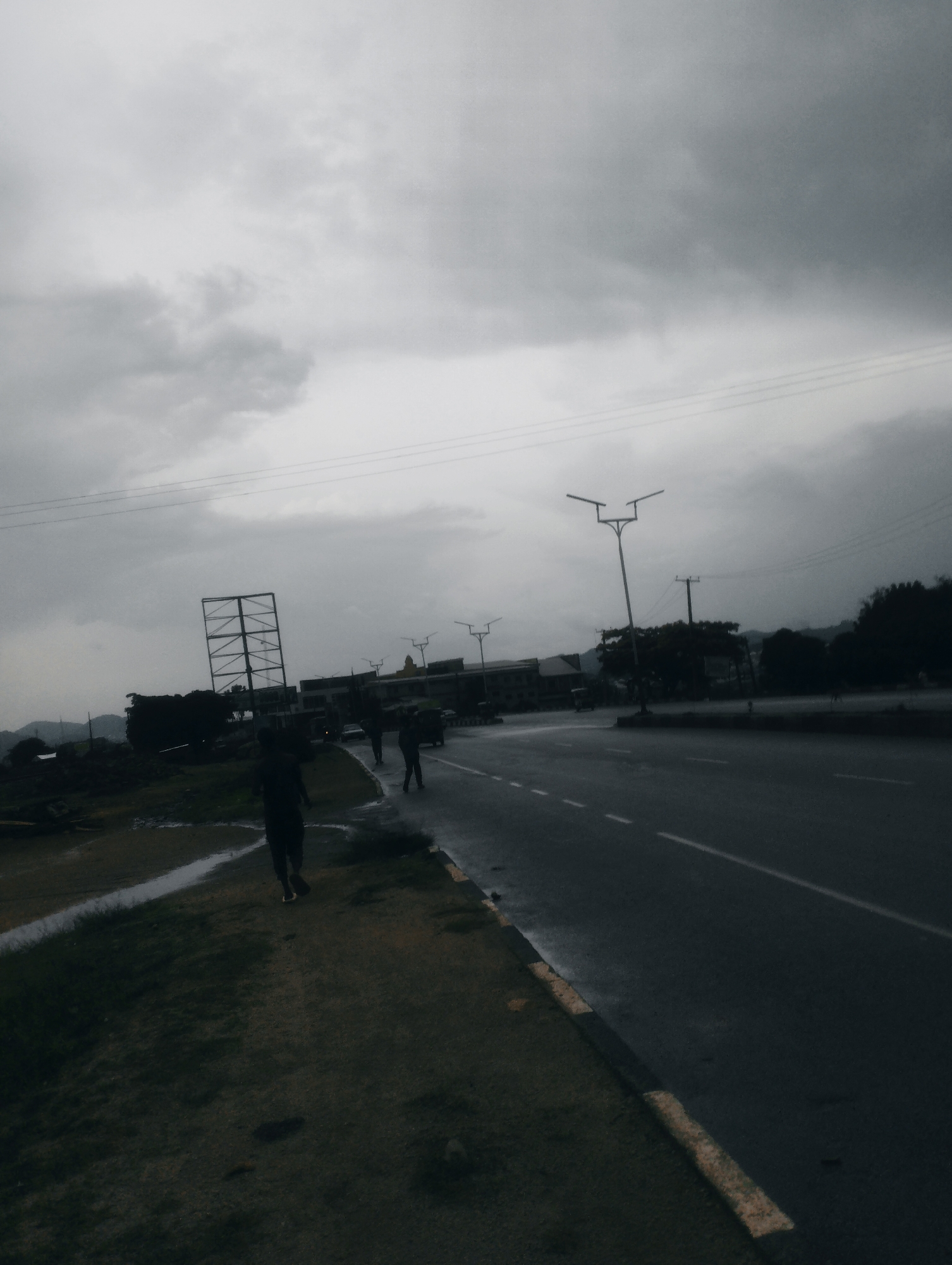 The image depicts a roadside scene in Jos, Nigeria, under an overcast sky. A dark, wet asphalt road with white dashed lines runs from the foreground towards the background, curving slightly to the right. On the left side of the road, a grassy verge slopes downwards, with some puddles and patches of wet ground visible. Several figures are walking along the grassy verge and on the edge of the road. One figure in the foreground is walking away from the camera on the grass. Further down the road, on the right side, several other figures are walking. In the background, there are buildings, trees, and streetlights. A large, metal scaffolding structure is visible on the left side of the road, near the background. The overall atmosphere is muted and somber, likely due to the cloudy weather.