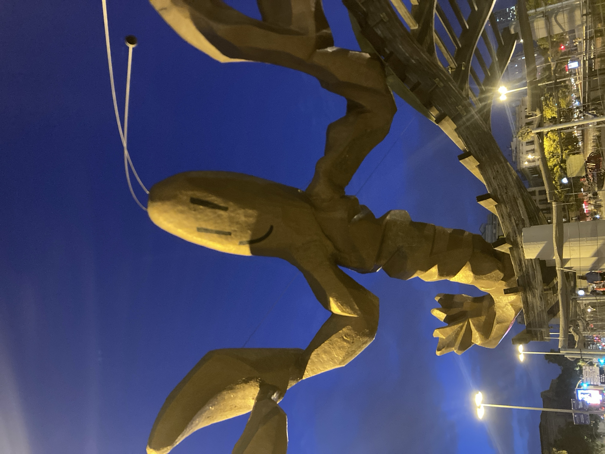 This image shows a large, golden sculpture of a lobster in Barcelona, Spain. The sculpture is located in a public space, possibly a plaza or park, and is visible at night. The sky is a deep blue, indicating it is either dusk or night. Streetlights and building lights are visible in the background, illuminating the surrounding urban environment. The lobster sculpture is the primary subject, with its large claws and segmented tail prominently displayed. The sculpture appears to be made of metal and has a stylized, somewhat abstract design. There are a few people visible in the background, walking or standing. The overall scene depicts an outdoor urban setting at night, with a prominent piece of public art as the focal point.