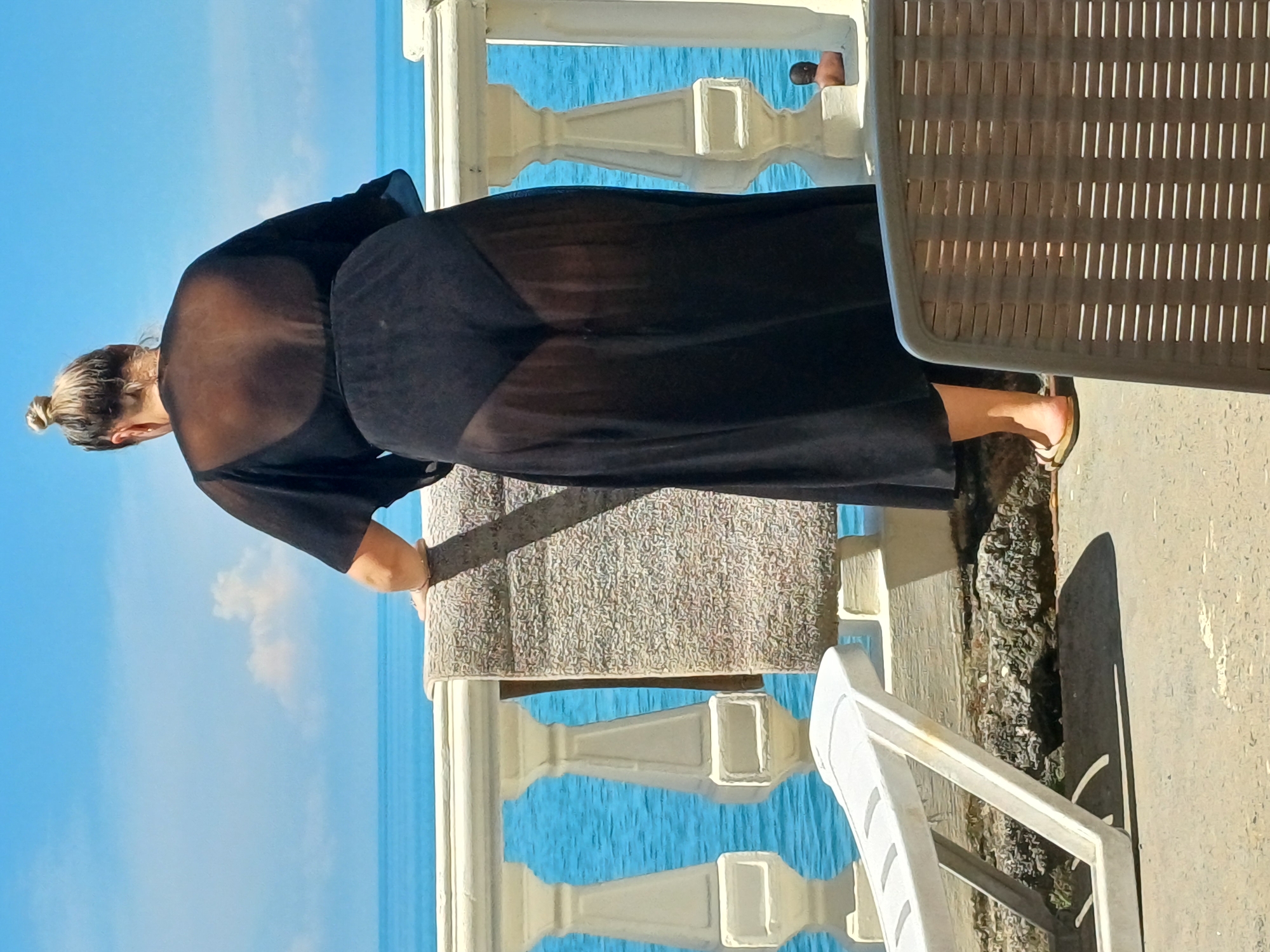 A woman, from behind, stands on a balcony overlooking the sea in Sochi, Russia. She is wearing a sheer black dress over a bikini bottom and a bra. She is holding a towel over the balcony railing. In the background, a man with no shirt on is visible near the water. A white lounge chair and a brown chair are also present on the balcony. The sky is blue with some clouds.