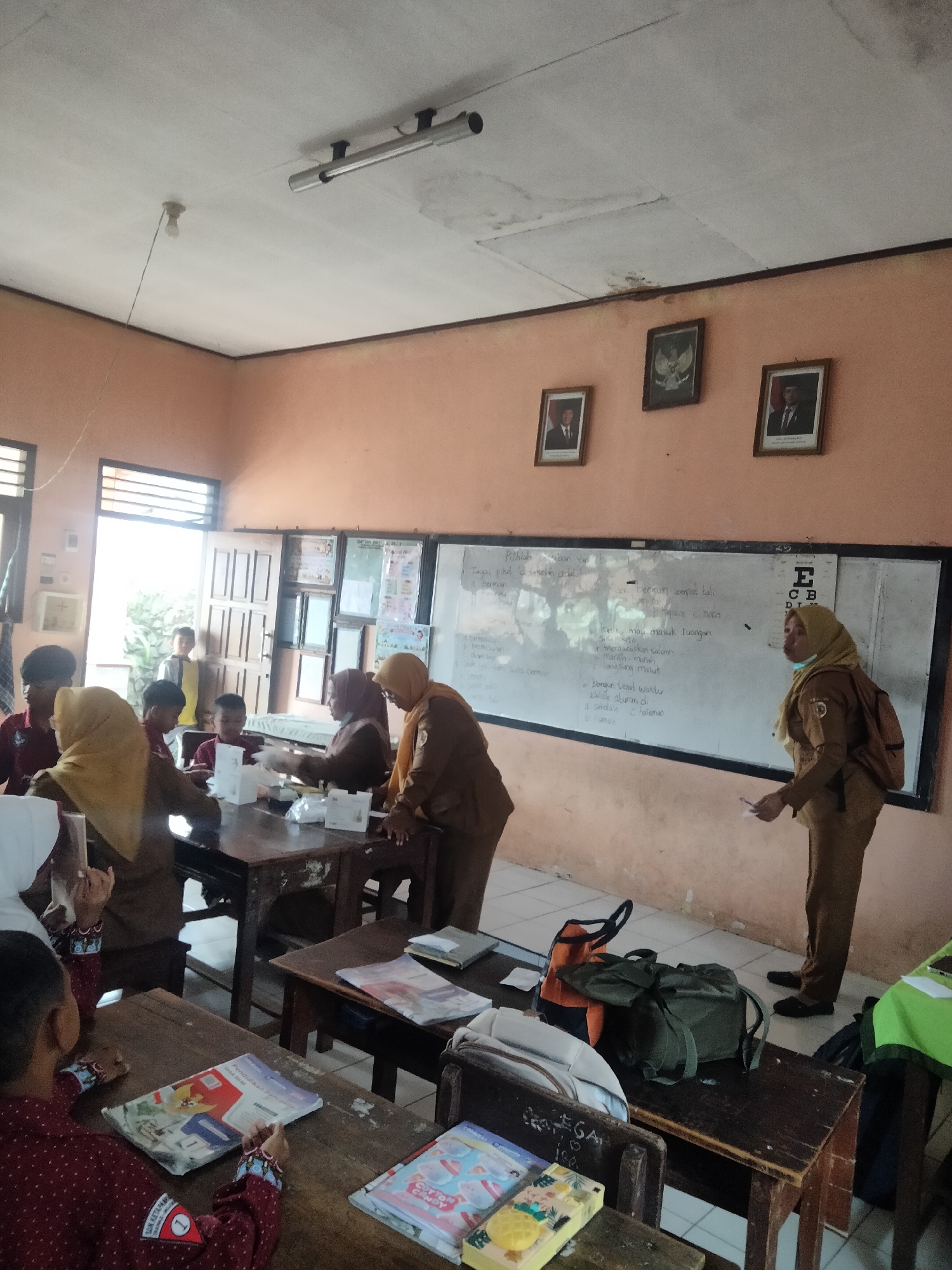 The image depicts a classroom setting in Tretes, Indonesia, with students and teachers engaged in an activity. Two female teachers, dressed in brown uniforms and headscarves, are interacting with a group of young students seated at wooden desks. One teacher stands near a whiteboard, holding papers, while the other is assisting students at a desk. Several students, some wearing uniforms, are gathered around a table with what appear to be educational materials. The classroom environment is characterized by simple wooden furniture, a whiteboard with writing, framed pictures on the wall, and fluorescent lighting. The overall scene suggests an informal learning or assessment session.