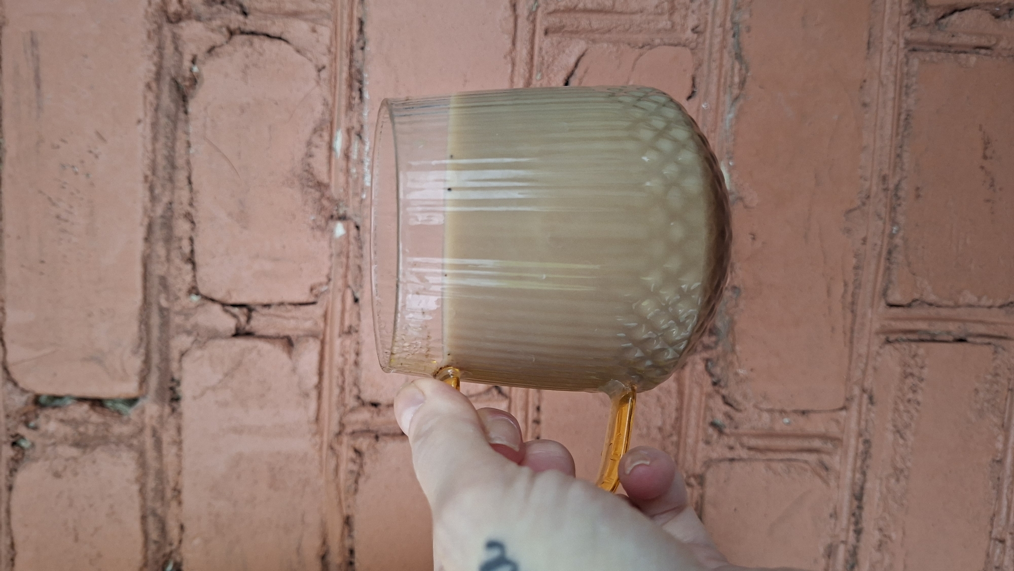 A hand with a tattoo on the wrist holds a clear glass mug filled with a light brown beverage. The mug has a textured pattern on the bottom half and vertical ridges on the top half, with an orange handle. The mug is positioned in front of a reddish-brown brick wall. The image was taken in the city of Myadzyel, Belarus.
