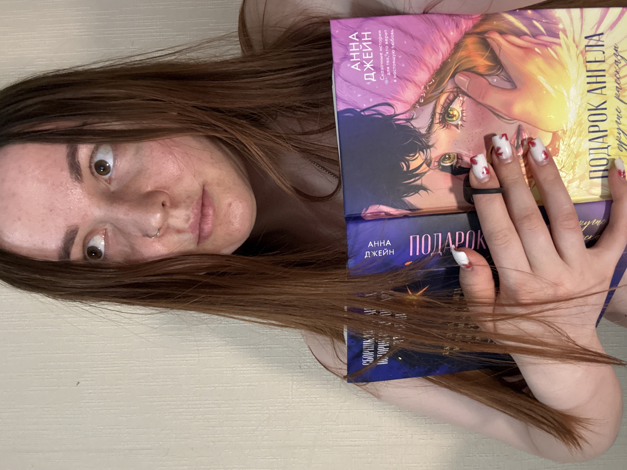 A young woman is depicted lying down, holding three books against her chest and face, in what appears to be an indoor setting in Izhevsk, Russia. The image is rotated 90 degrees, showing her head to the left and body extending to the right. She has long, straight brown hair that is spread out around her head and over her left arm. Her eyes are wide, and she has a slightly open mouth, conveying a surprised or playful expression. A silver nose ring is visible on her right nostril.
Her hands are holding the books, with her fingers adorned with white nails featuring distinctive red, possibly heart or blood-splatter, designs. A black rubber band is wrapped around one of her fingers. The books are stacked, and their covers display colorful illustrations and text in Russian.
Visible text on the books includes the author's name "АННА ДЖЕЙН" (Anna Jane). One book's cover reads "Сказочные истории для тех, кто верит в настоящую любовь" (Fairy tales for those who believe in true love). Another cover states "ПОДАРОК АНГЕЛА другие рассказы" (Angel's Gift other stories), and partially visible text on a third book suggests "СБОРНИК ИСТОРИЙ" (Collection of Stories). The background is a plain, light-colored surface, possibly a wall or floor.