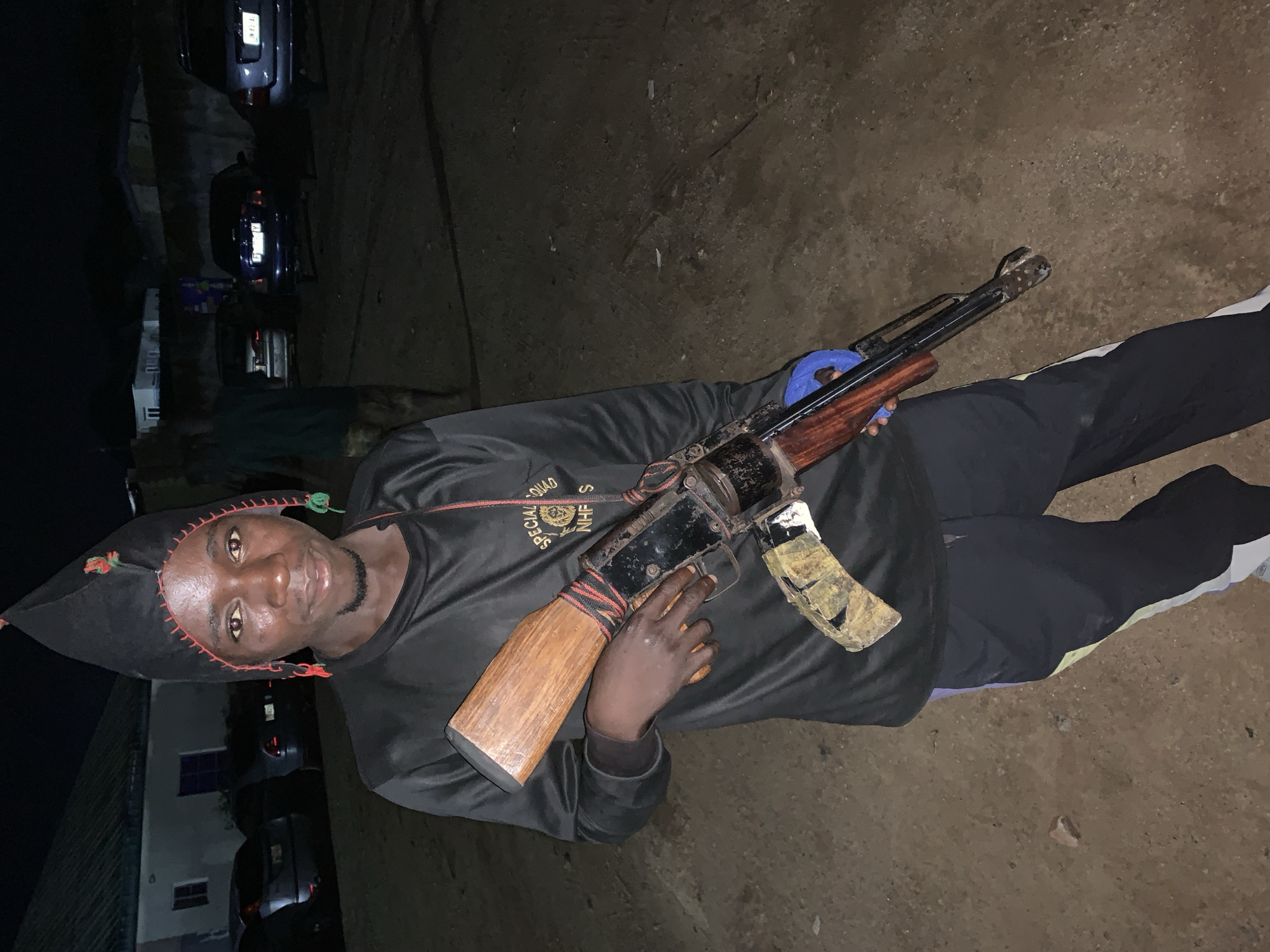 A man is holding a rifle while standing outdoors. He is wearing a black jester hat, a black long-sleeved shirt with red and white lettering, and black pants with a white stripe. The rifle appears to be an AK-47 variant with a wooden stock and pistol grip. The magazine has some camo tape on it. Behind him is a building, and a car can be seen in the background. The ground is dirt. Based on the request, this is in Kabong, Nigeria.
