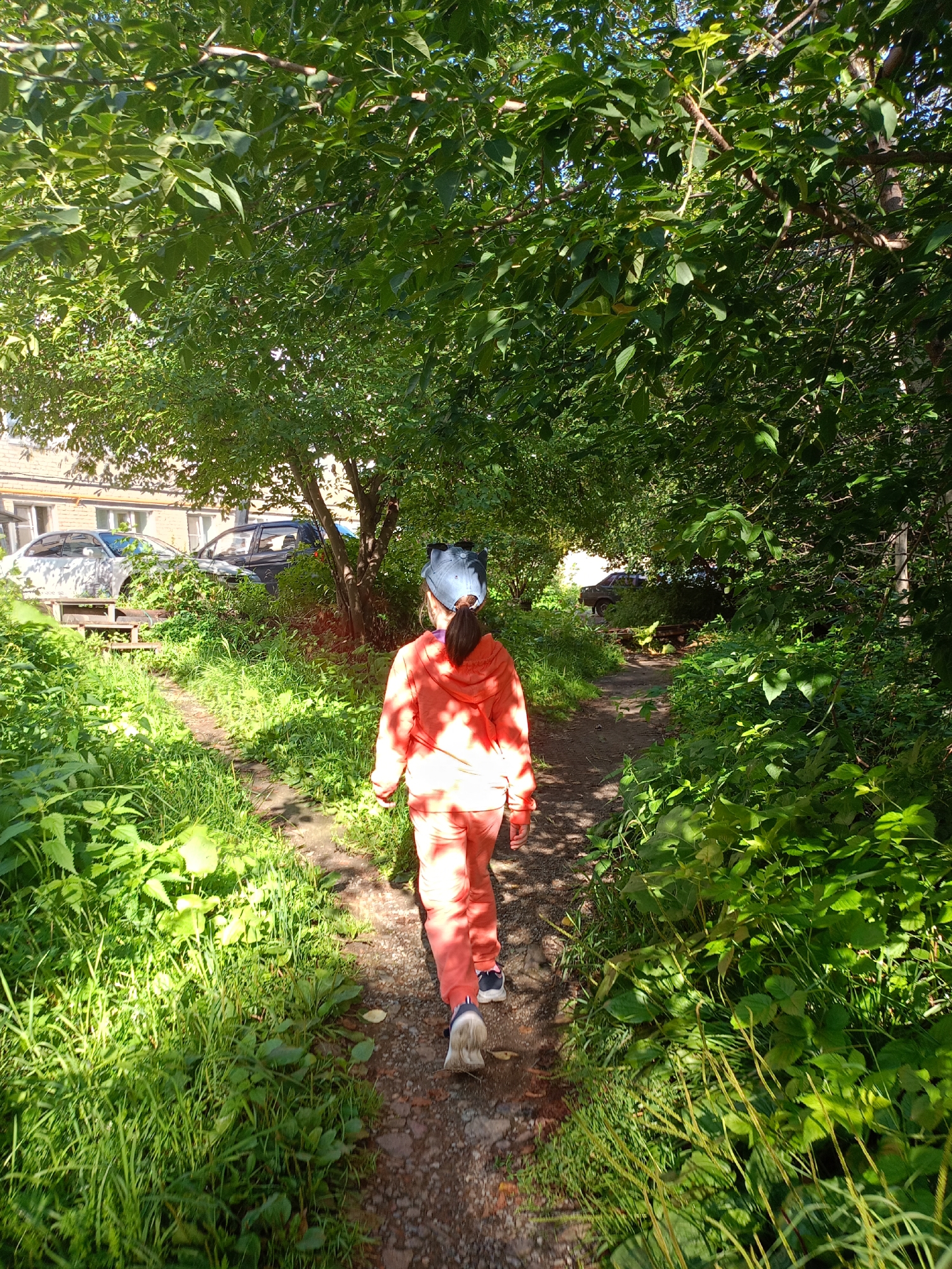A young person wearing a bright orange hooded jacket and matching pants, along with a blue baseball cap, walks away from the camera down a dirt path. The path is flanked by lush green vegetation and is shaded by the canopy of trees overhead. In the background, through a break in the trees, a brick building with several windows is visible, as well as two parked cars. The scene appears to be in an urban or suburban area of Zlatoust, Russia, likely a courtyard or park area. The lighting suggests it is daytime, with sunlight filtering through the leaves.