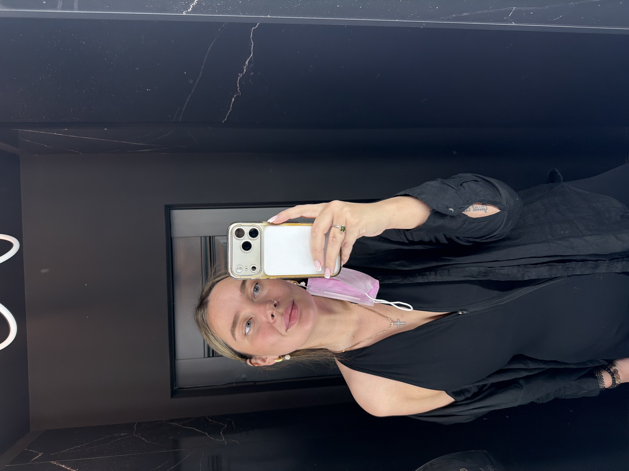 A young woman is captured in a mirror selfie, standing upright in what appears to be a modern indoor setting, possibly in Moscow, Russia. She holds a white smartphone with a multi-lens camera system to take the photo, partially obscuring her face.
The woman has light-colored hair, which frames her face, and is wearing a black sleeveless top with a zipper down the front, layered under a loose black long-sleeved shirt or jacket. A light pink disposable face mask hangs from her left ear. She wears a silver-toned necklace with a cross pendant, pearl earrings, and a small stud nose piercing. A ring with a green stone is visible on her right hand, and a multi-strand bracelet adorns her left wrist. On her right forearm, a tattoo with text in a non-Latin script is partially visible. Her expression is neutral, with her lips slightly parted.
The background consists of dark, reflective surfaces, resembling black marble or a similar material with fine white veining, suggesting a sleek and contemporary interior. A prominent white, illuminated, abstract light fixture is visible on the left side of the reflection. The overall scene conveys a stylish and modern environment.