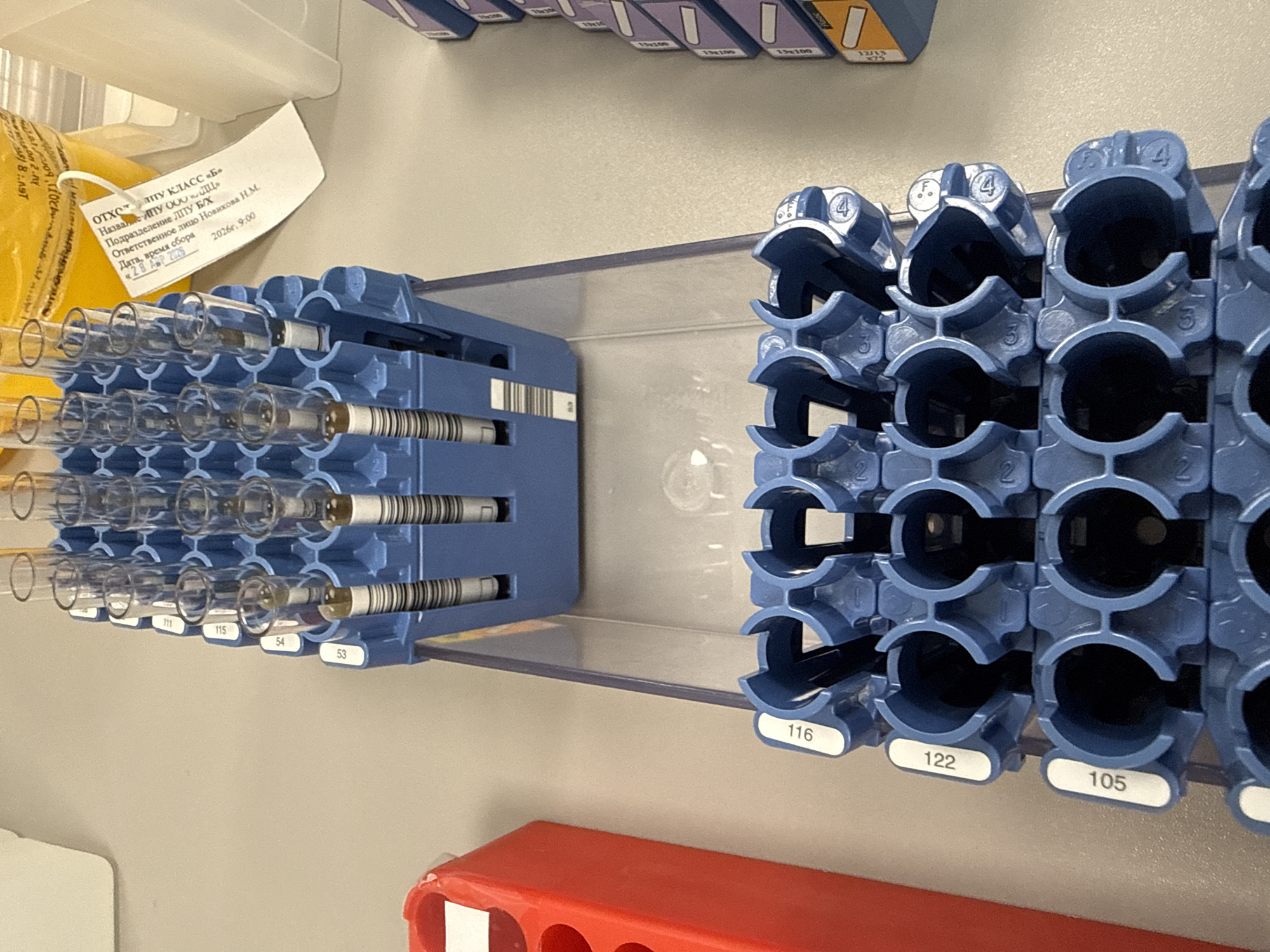 The image depicts a close-up of laboratory equipment, likely within a medical or research facility. The primary subjects are blue plastic racks designed for holding test tubes. On the left, one rack is filled with clear glass test tubes, each containing a liquid and secured with a cap and barcode label. Numbers such as "711", "115", "54", and "53" are visible on this rack. To the right, a similar blue rack, mostly empty, features individual slots marked with numbers like "2", "3", and "4", and labels "116", "122", and "105" on its front.
A paper label, written in Russian, is attached to a yellow bag or container in the upper left. The text reads: "ОТХОТ АПУ КЛАСС «Б»" (Waste APU Class "B"), "Название ИПУ ООО «ДЦ" (Name IPU OOO "DC"), "Подразделение ЛІТУ Б/Х" (Department LPU B/X), and "Ответственное лицо Новикова Н.М." (Responsible person Novikova N.M.). It also indicates the "Дата, время сбора" (Date, time of collection) as "28 АПР 2026" (28 April 2026) at "9:00" (9:00 AM). This suggests the activity involves the organization or storage of medical samples, possibly waste, collected on that specific date and time.
In the background, out-of-focus containers bear text like "13x100," likely indicating dimensions. A red plastic tray is visible at the bottom. The bright lighting suggests an indoor lab environment. The Cyrillic text on the label, combined with the Moscow location context, places the scene in a Russian laboratory or medical institution.