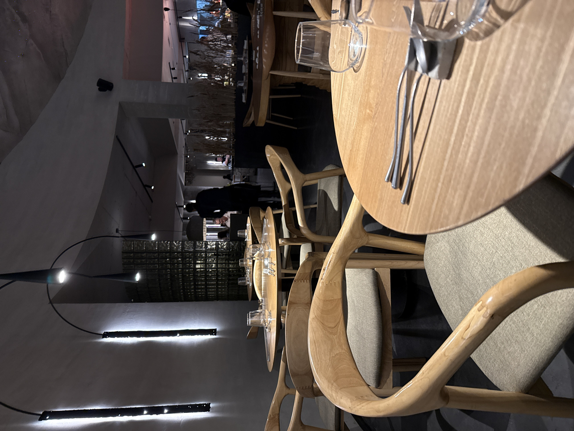 This image captures the interior of a modern and elegantly designed restaurant or dining establishment, likely during the evening due to the dim, atmospheric lighting. The setting suggests an upscale dining experience.
In the foreground, several round dining tables made of light-colored wood are visible, each meticulously set with clear glasses and a set of polished silver cutlery. The chairs accompanying these tables are also crafted from light wood, featuring distinctive curved, organic designs and light grey fabric seats.
The scene extends into the midground, revealing more dining tables and chairs arranged throughout the space. In the background, indistinct figures, including one person in dark attire, are moving within the restaurant, suggesting activity. A dark area in the far background features decorative elements that resemble tall, dried grasses or reeds, illuminated from behind, adding to the sophisticated ambiance.
Notable details include various lighting fixtures: long, horizontal light strips are mounted on a light-colored wall, emitting a soft white glow, and small, black, cylindrical spotlights are visible on pillars or walls. A grid-like screen or decorative partition is also present in the middle ground, adding textural interest. The overall aesthetic emphasizes natural materials and clean lines. No discernible text is visible within the image. The location context provided is Krasnoyarsk, Russia.
