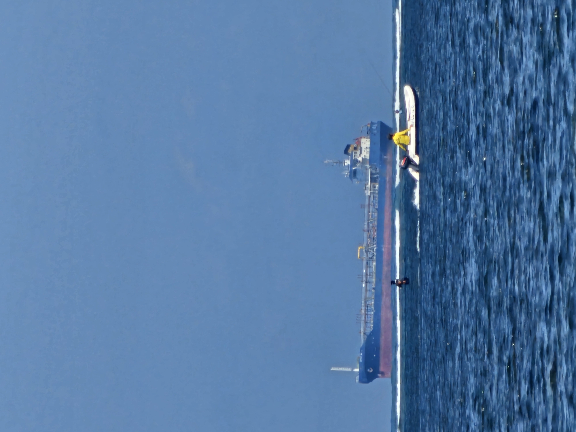 A large blue and red cargo ship is on the horizon under a clear blue sky. In the foreground, a small white boat with a person in a yellow shirt is moving to the left. Another person is visible in the water near the boat. The water is choppy and blue. The scene appears to be a coastal area, possibly near the city of Denpasar, Indonesia, given the presence of the cargo ship and boats.