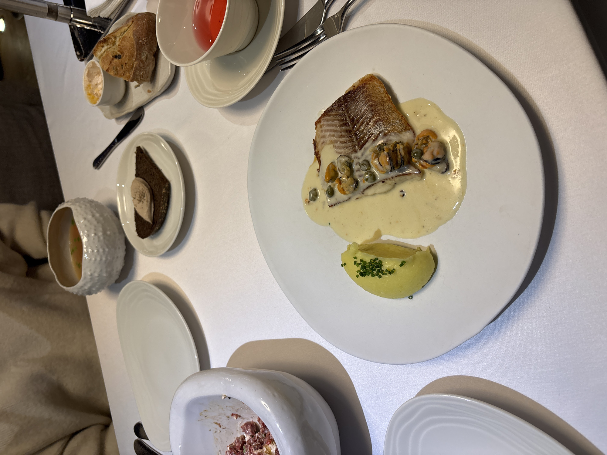 An interior restaurant scene in Krasnoyarsk, Russia, displays a white-tablecloth-covered dining table set with multiple courses. In the foreground, a white round plate presents a pan-seared fish fillet topped with a creamy sauce, mussels, and capers, accompanied by a quenelle of chive-garnished mashed potatoes. To the immediate left, a white ceramic mortar contains a reddish, finely minced substance, indicating partial consumption. Further left, an empty elongated oval white plate and a textured white bowl holding an amber broth with green specks are visible. On the right, a small white plate displays a slice of dark bread with a light-colored spread. Adjacent, a small rectangular tray holds a piece of crusty bread and a white ramekin with a yellowish spread. A white teacup containing a red liquid rests on a saucer in the lower-right. Silverware, including a fork and knife, is positioned to the right of the main plate. The torso and arms of a person in a light-colored garment are partially visible in the upper-left background, indicating a seated diner.