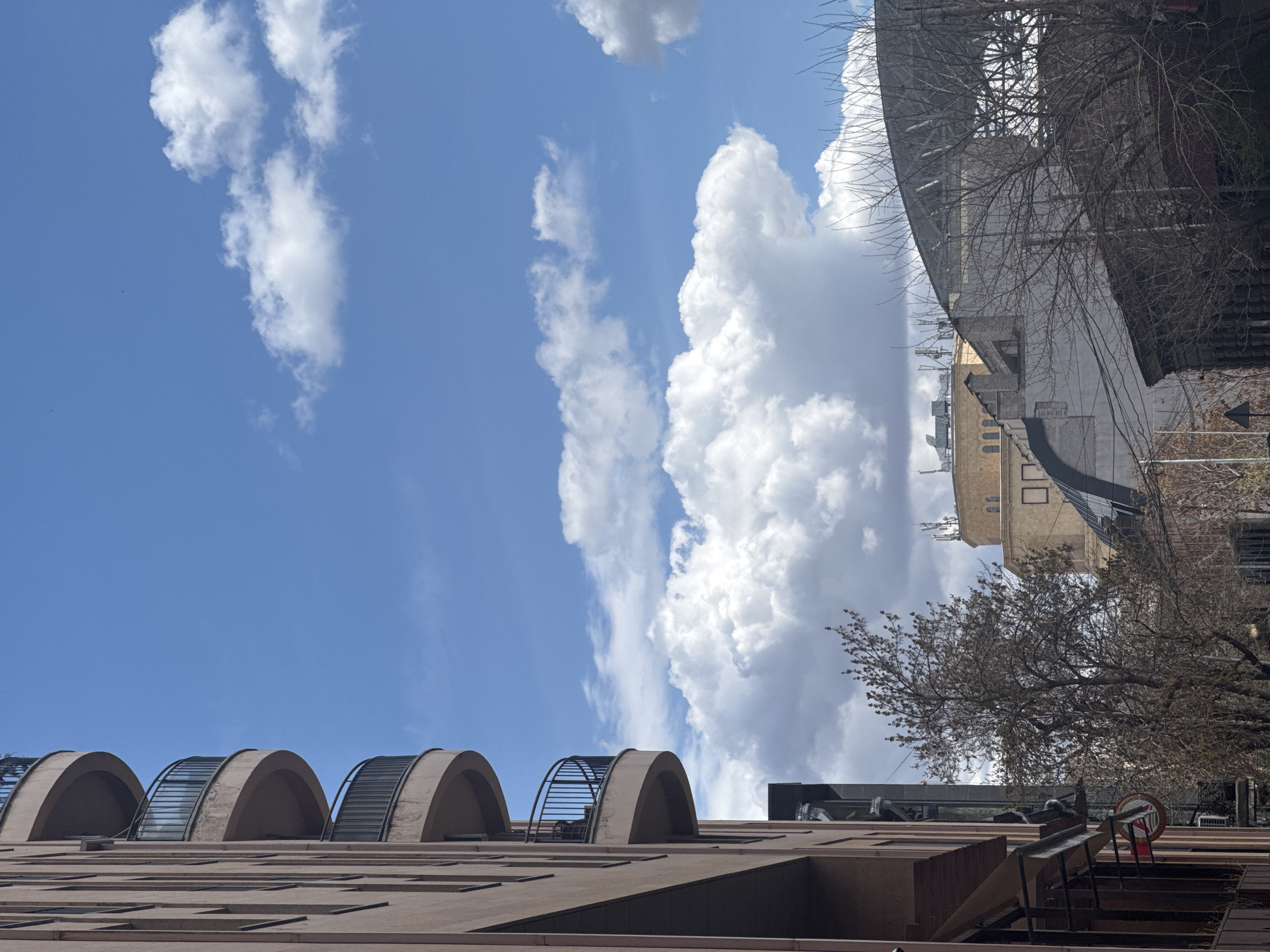 The image depicts a low-angle view of buildings in Yerevan, Armenia, under a partly cloudy sky. The sky is predominantly blue with scattered cumulus clouds. The lower portion of the image shows a building with a facade featuring arched windows or alcoves with metal railings. To the right, a taller building complex is visible, displaying a mixture of architectural styles and textures, including a curved metal structure possibly a roof or architectural feature, and bare tree branches silhouetted against the sky. A red and white prohibition sign is visible in the foreground. The overall impression is of a modern urban environment with architectural details characteristic of Yerevan.