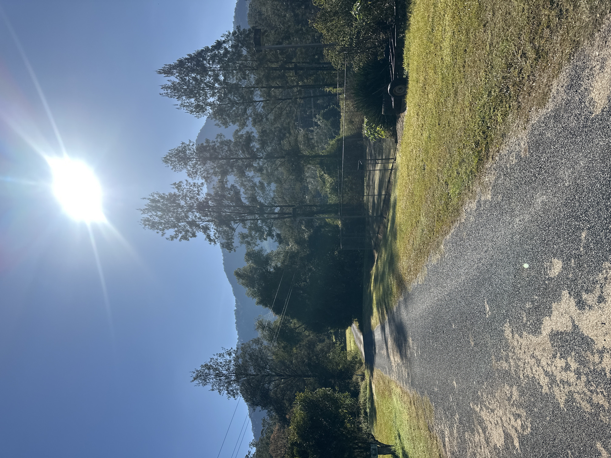 A bright, sunny day illuminates a gravel driveway leading into a rural landscape. The sun, a brilliant orb in the clear blue sky, casts a lens flare effect across the top of the image, with its rays stretching outwards. Tall, slender trees with dense foliage line the right side of the driveway, interspersed with shadows from the overhead sun. Beyond the trees, a chain-link fence encloses an area that appears to be a tennis court, its surface visible through the mesh. A dark, two-wheeled trailer with a metal frame is parked on the grassy area next to the fence. In the distance, a hazy mountain range rises, its slopes covered in a thick carpet of trees. The foreground is dominated by the textured grey gravel of the driveway, with patches of light and shadow creating a dappled effect. The overall impression is one of a peaceful, sun-drenched rural setting.