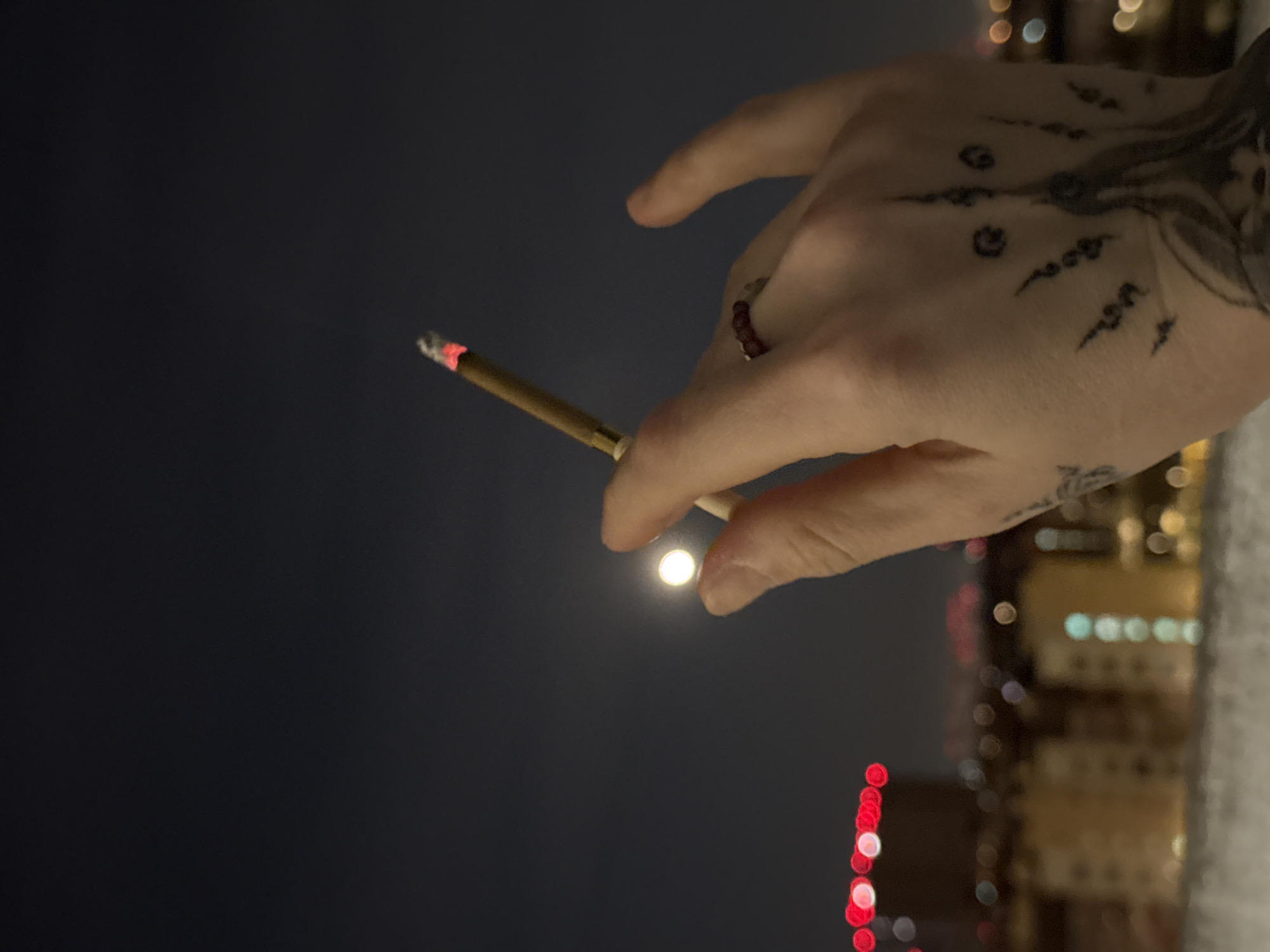 A hand holding a lit cigarette or joint is in the foreground. The hand has several tattoos and a beaded ring on one finger. The background shows a dark sky with a small, bright light source appearing to be a full moon between the index and middle fingers of the hand. Out-of-focus city lights, including red and yellow bokeh, are also visible in the lower right of the frame. The specific location in Moscow, Russia cannot be determined from the information in the image.