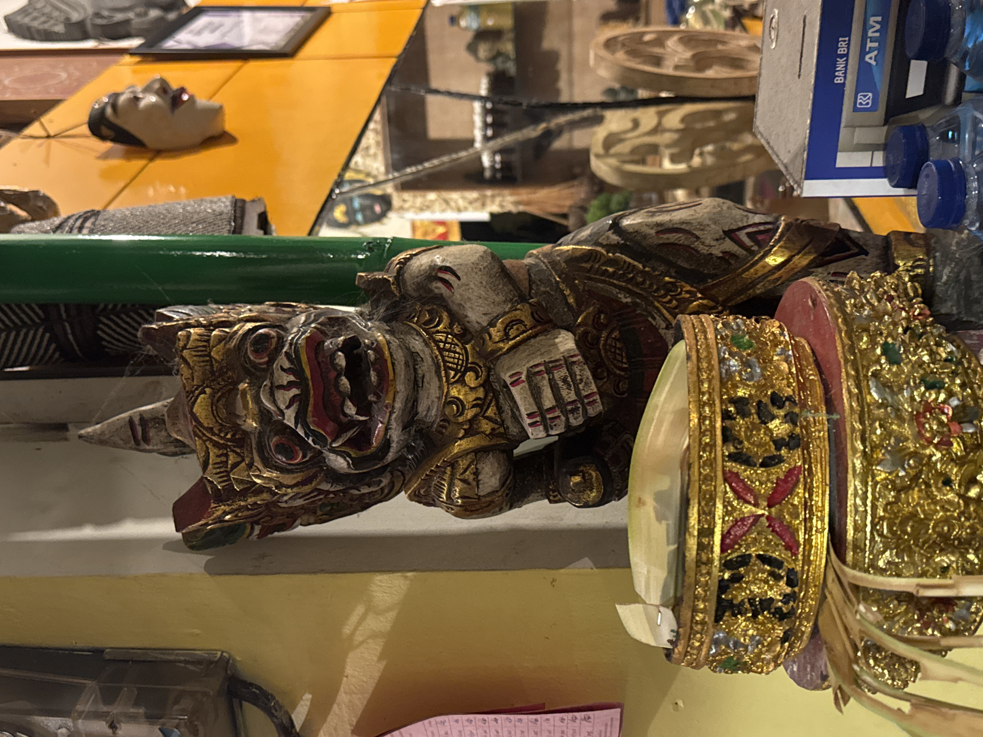 The image shows a close-up of a carved wooden demon-like figure with gold accents. It has prominent eyes, horns, and a red-painted mouth. The figure is partially obscured by ornate, gold-colored bracelets with gem-like embellishments and red and green accents. A green bar runs across the top of the figure. In the background, various objects are visible, including a yellow surface, a face-like mask, circular decorative items, a Bank BRI ATM box, and water bottles. There is a shelf beneath the figure. The location is Kuta Selatan, Indonesia.
