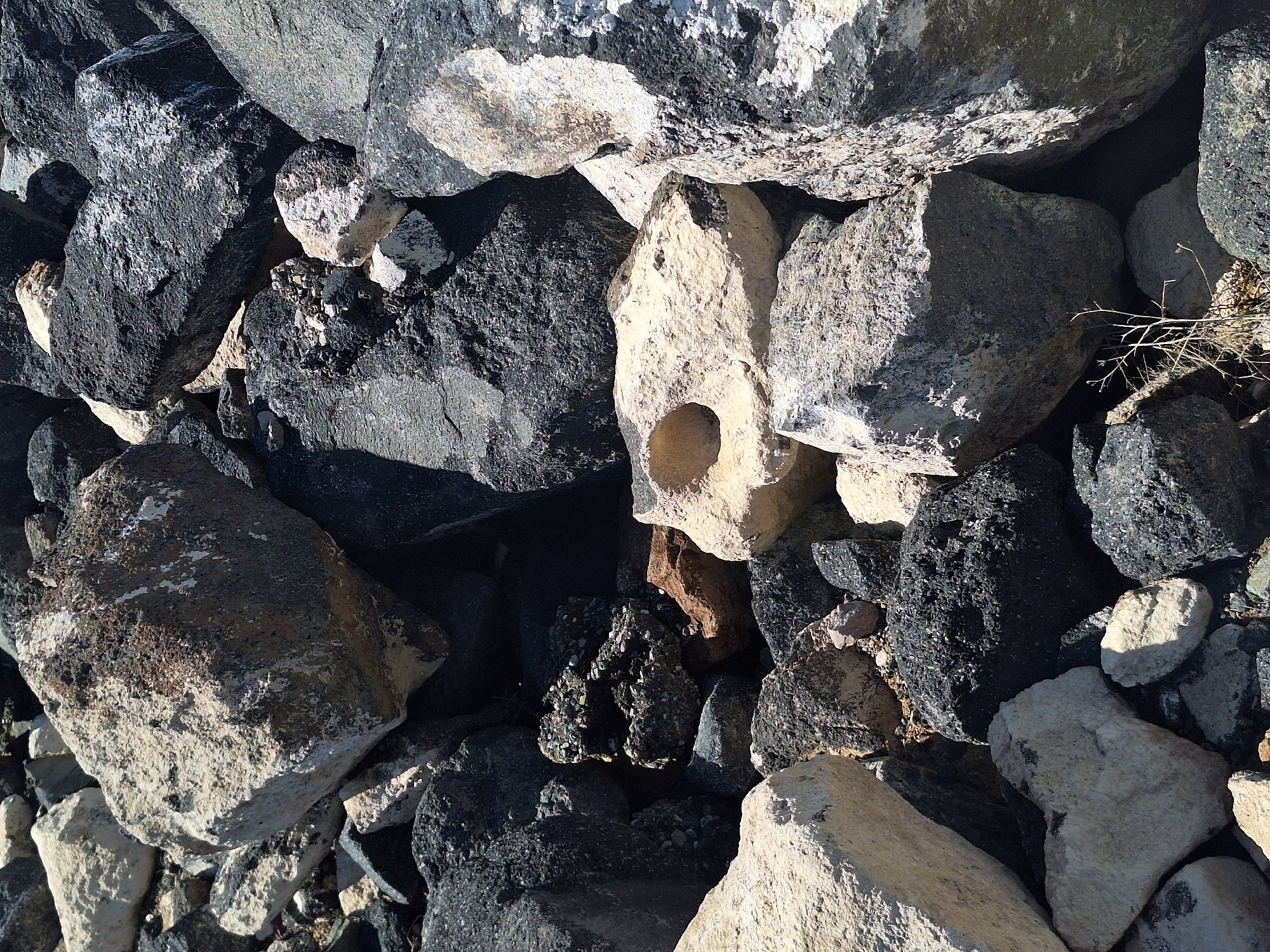 The image shows a pile of rocks of various sizes, shapes, and colors. The rocks are mostly gray and black, with some lighter-colored stones mixed in. One of the lighter-colored rocks has a distinct hole in it. The scene appears to be located in a natural environment, such as a rocky hillside or a riverbed. There are no people or animals visible in the image. The image was captured in Aniavan, Armenia.