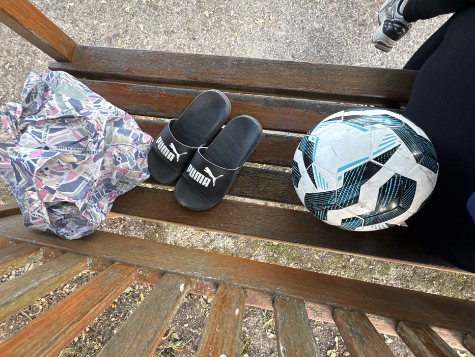 The image presents a downward-angled view of objects resting on a weathered wooden park bench in Conflans-Sainte-Honorine, France. From top to bottom, a crumpled multi-colored fabric item, possibly a shirt or scarf, is visible. It features a map-like pattern with text elements including "AV DES CHAMPS," "KLEBER," and "PARIS." Below this, a pair of black Puma-branded slides with white logos are placed side-by-side. Further down, a white, black, and light blue Adidas soccer ball with visible scuff marks and dirt occupies a section of the bench. The lower right corner of the frame includes a portion of a person's lap, covered by dark blue or black fabric, and the front of a silver, black, and white athletic shoe, indicating a seated individual. The ground adjacent to the bench consists of light-colored gravel and sparse, dried vegetation. The scene is illuminated by natural daylight.
