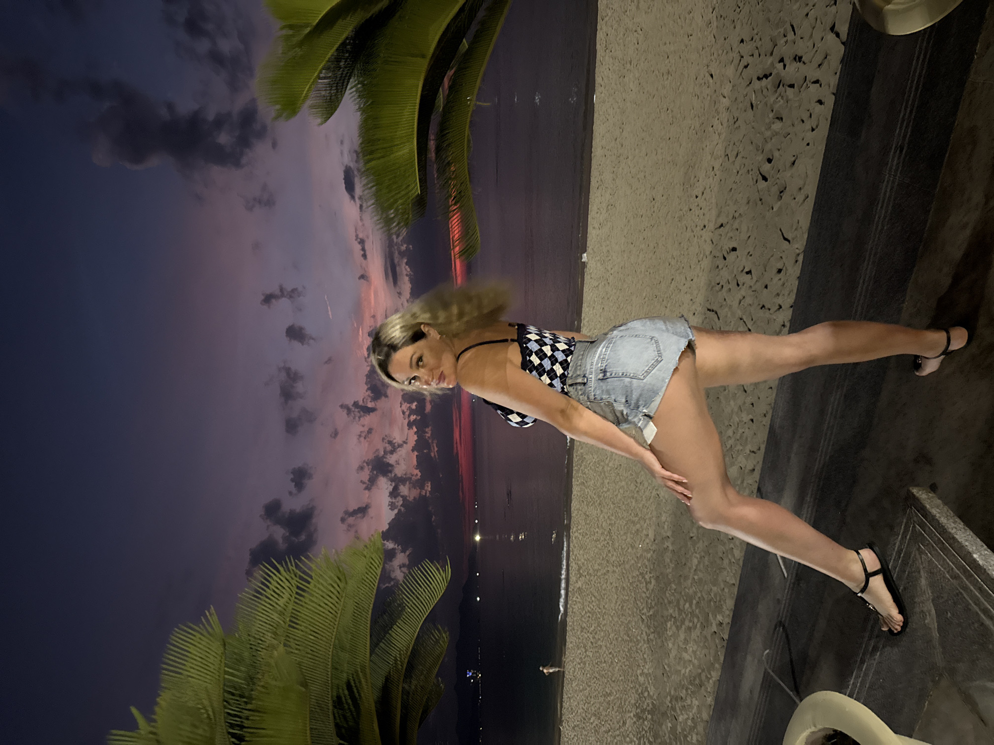 A woman is posing on a concrete ledge in Nha Trang Ward, Vietnam. She is wearing a checkered top and jean shorts, with black sandals. The background consists of a beach at dusk, with palm trees framing the scene. The sky transitions from dark purple to pink. The woman is leaning on the ledge, looking towards the camera. The beach shows lights from distant boats or structures.