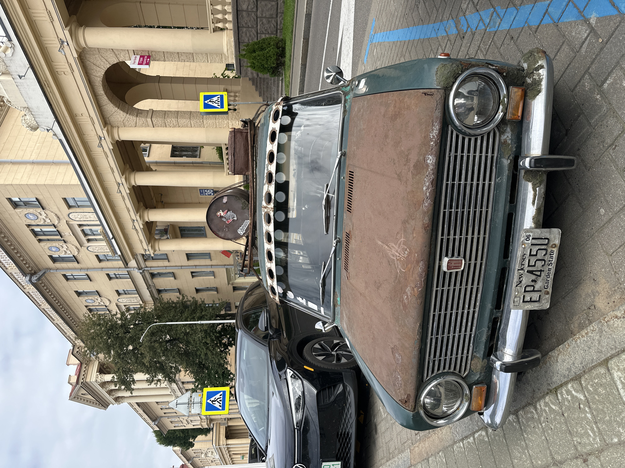 A vintage, rusty green car is parked on a paved street in Minsk, Belarus, in front of a grand, cream-colored building with classical architecture. The car has a New Jersey license plate that reads "EP-455U GARDEN STATE". It also has items loaded on its roof, including what appears to be a metal drum and a small wooden crate. A black Volkswagen ID.4 is parked to the left of the vintage car. Yellow pedestrian crossing signs are visible on both sides of the street, with a crosswalk marked with blue lines in the foreground. The sky is overcast.