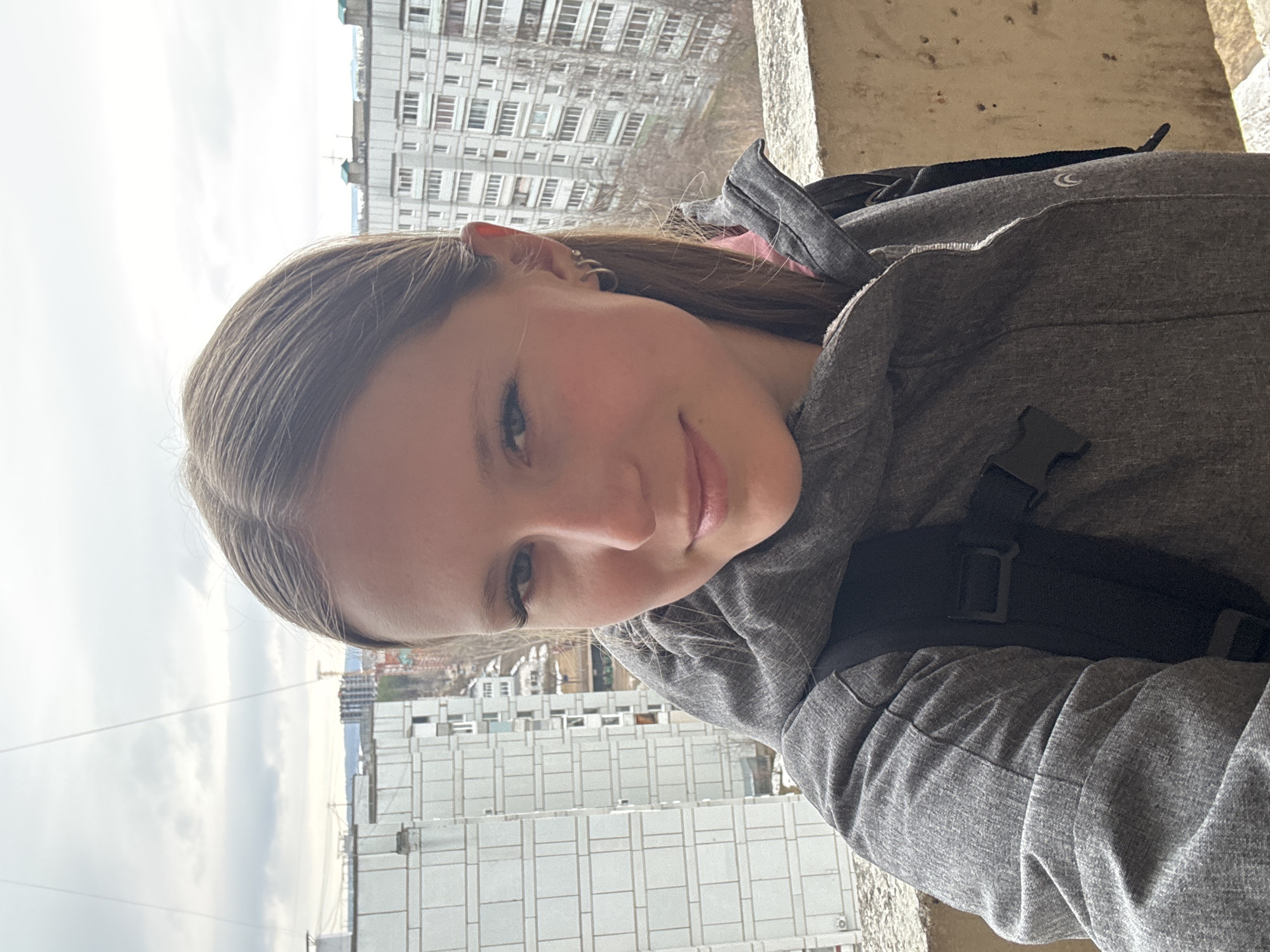 A young woman with light brown hair and blue eyes is taking a selfie. She is wearing a gray hooded jacket and a black backpack. The background shows a cityscape, with apartment buildings. Based on the architectural style and the presence of typical Soviet-era apartment blocks, the location is identified as Chaikovsky, Russia.