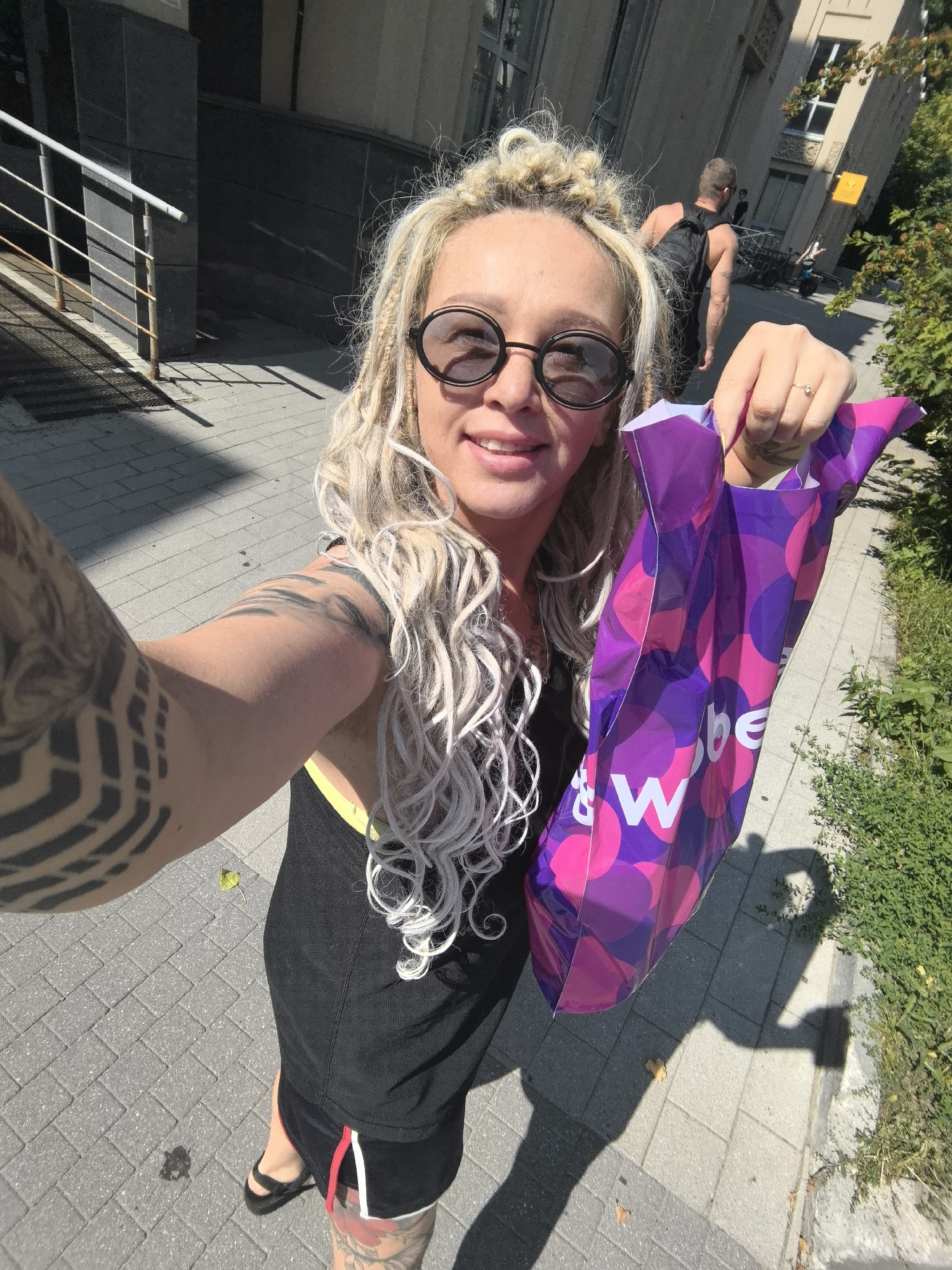 A woman is taking a selfie outdoors in what appears to be a city. She has blonde, braided hair and is wearing sunglasses, a black tank top, and black shorts. She is holding a purple and pink patterned shopping bag with the letters "W" and "&" visible. A man with a backpack is walking away in the background, and trees and buildings are visible. The ground is paved with gray bricks. The scene is illuminated by sunlight, casting shadows.