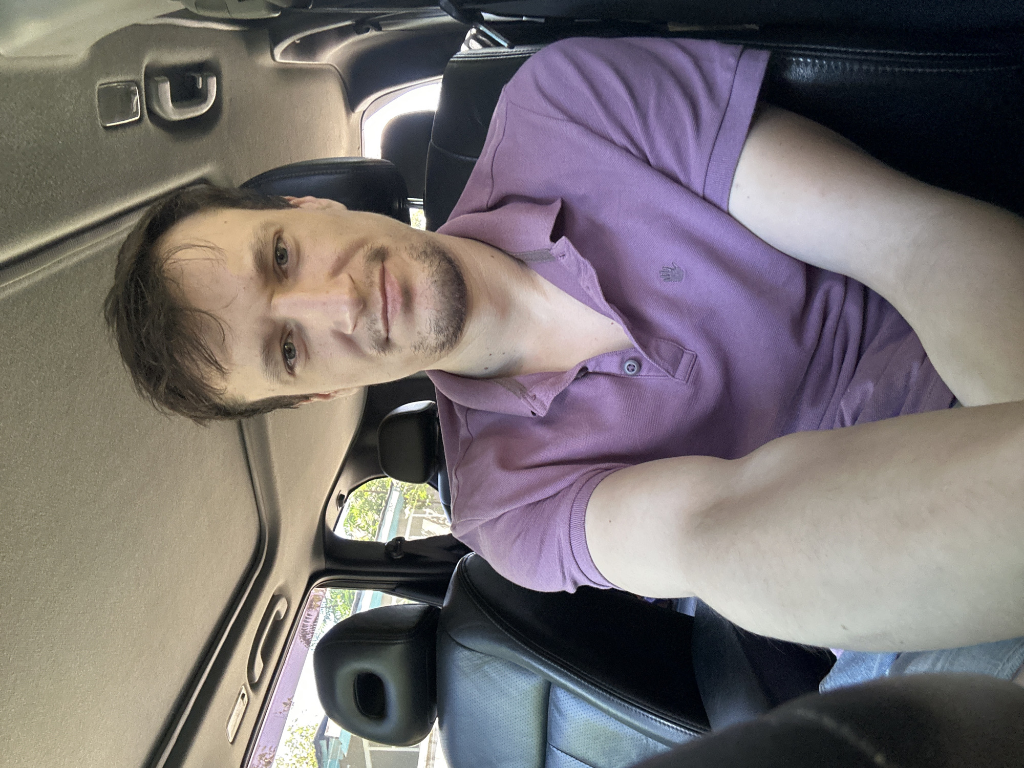 A man is sitting in the driver's seat of a car, looking at the camera. He is wearing a purple polo shirt. The interior of the car is visible, including black leather seats and a sunroof. The man has short brown hair and a light beard. The background shows trees and buildings, suggesting an outdoor setting within a city. The lighting indicates it is daytime. The location is identified as Makiv, Ukraine.