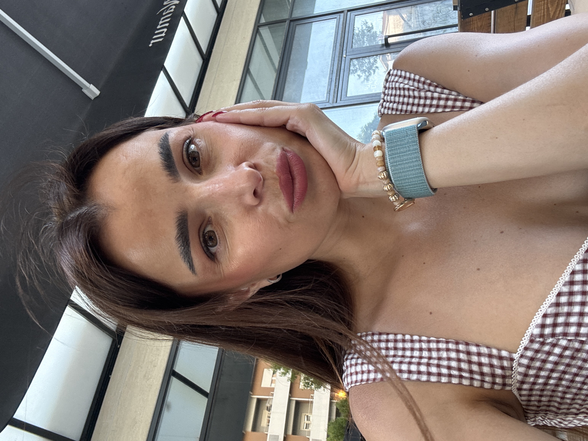 A woman is pictured in a close-up selfie, taken outdoors. She is leaning her head on her hand, with her fingers resting on her cheek and jawline. Her hair is long and brown, framing her face. She is wearing a brown and white checkered top. A watch with a teal band and beaded bracelets adorn her wrist. The background shows a building with multiple floors and windows, suggesting an urban environment. The overall lighting indicates it is daytime. Based on the distinct architectural style of the building in the background, this location appears to be Podgorica, Montenegro.