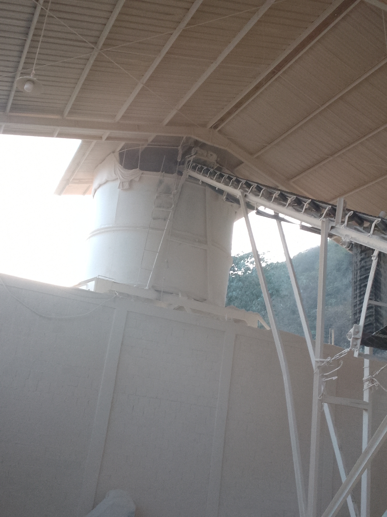 The image depicts an industrial setting, likely a processing plant, in Gunungmasigit, Indonesia. The foreground is dominated by a large, light-colored structure, possibly a building or silo, with a textured surface resembling concrete or plaster. Above and to the right, a conveyor belt system, also light-colored, is visible, supported by a metal frame. This system appears to be in operation or ready for operation, with visible components like rollers and a belt. In the background, a tall, cylindrical structure, likely a silo or storage tank, is partially covered with a light-colored tarp or sheeting. A ladder extends up the side of this cylinder, suggesting access for maintenance or operation. The overall environment is dusty, with a light-colored residue coating many surfaces, consistent with material processing. The roof structure above is composed of corrugated metal panels supported by a metal framework, with a single light fixture hanging from the ceiling. Beyond the industrial structures, lush green foliage and a hint of a hill or mountain are visible, indicating the natural environment surrounding the industrial site. The lighting suggests it is daytime, with the sun partially obscuring some areas of the image with its brightness. No people are visible in the image.