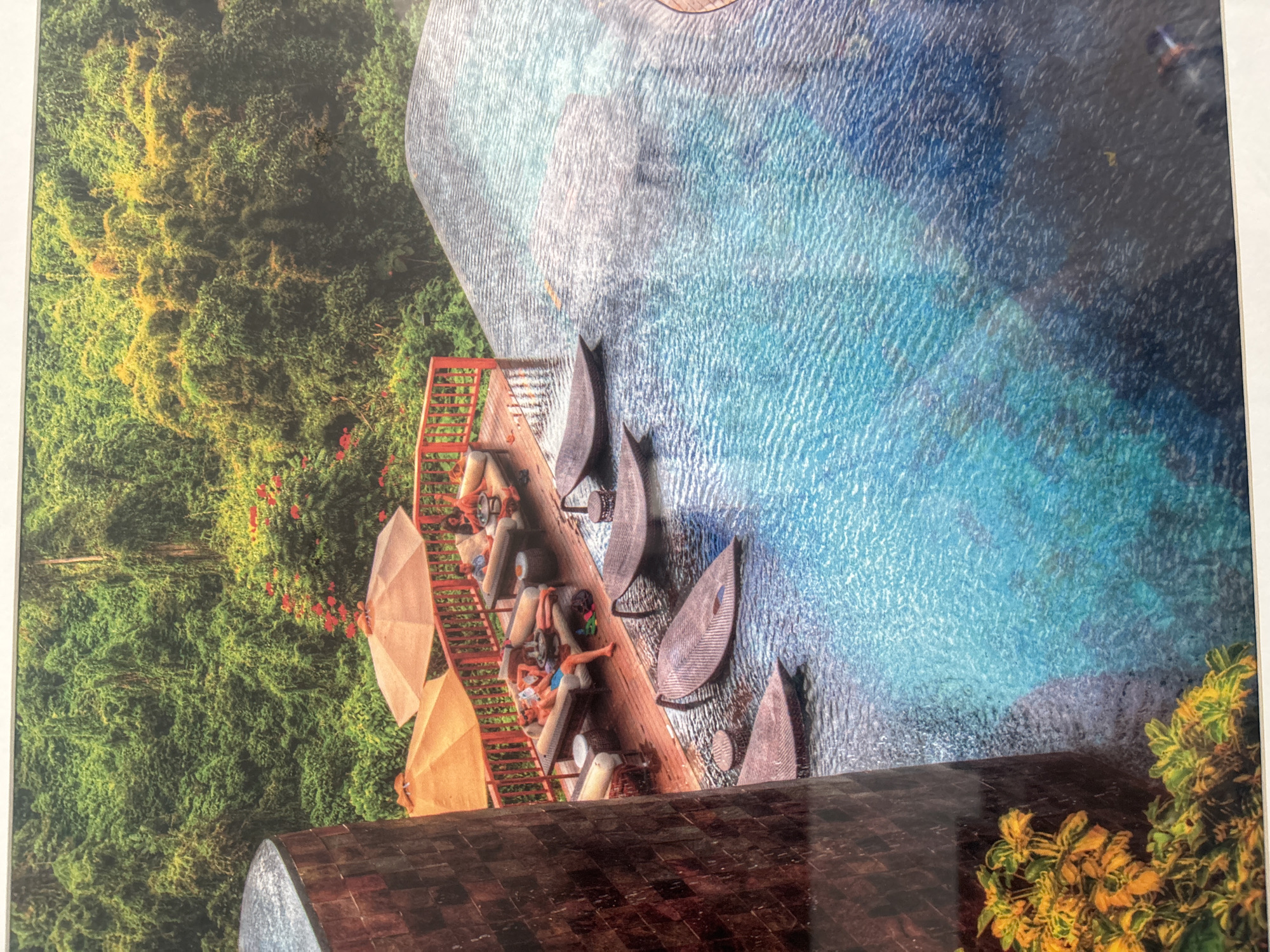 The image depicts an infinity pool overlooking a dense, green jungle landscape. The pool features several lounge chairs, some partially submerged in the water, shaped like elongated leaves. On a wooden deck adjacent to the pool, two orange umbrellas provide shade over lounge seating areas. Several individuals are visible relaxing on these loungers. In the foreground, a textured, cylindrical structure, possibly a pillar or column, partially obstructs the view. Lush vegetation with yellow and green leaves is situated at the bottom left of the frame. The overall scene conveys a sense of tropical luxury and relaxation. The provided image does not contain any elements that indicate the location is Dubai, United Arab Emirates.