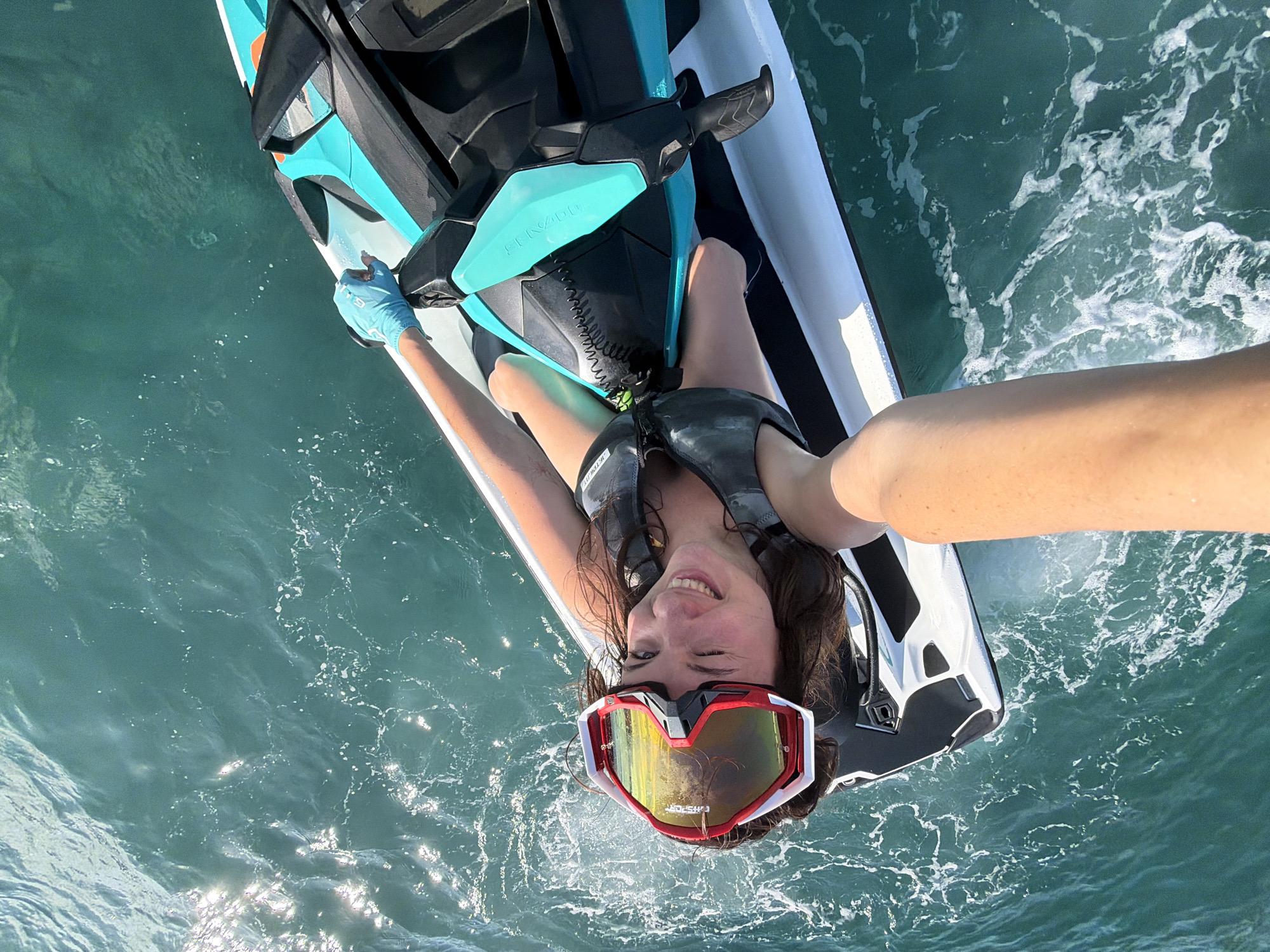 In Nusa Penida, Indonesia, a person is captured from an overhead perspective riding a jet ski on a sun-drenched, turquoise ocean. The individual, wearing a life vest and goggles, smiles joyfully at the camera, which appears to be held by another person or mounted to the front of the jet ski. The jet ski, a modern model with teal accents, is cutting through the water, creating white foam. Sunlight glints off the surface of the sea, highlighting the movement and clarity of the water. The scene is dynamic and suggests an exhilarating experience of recreational water sports.
