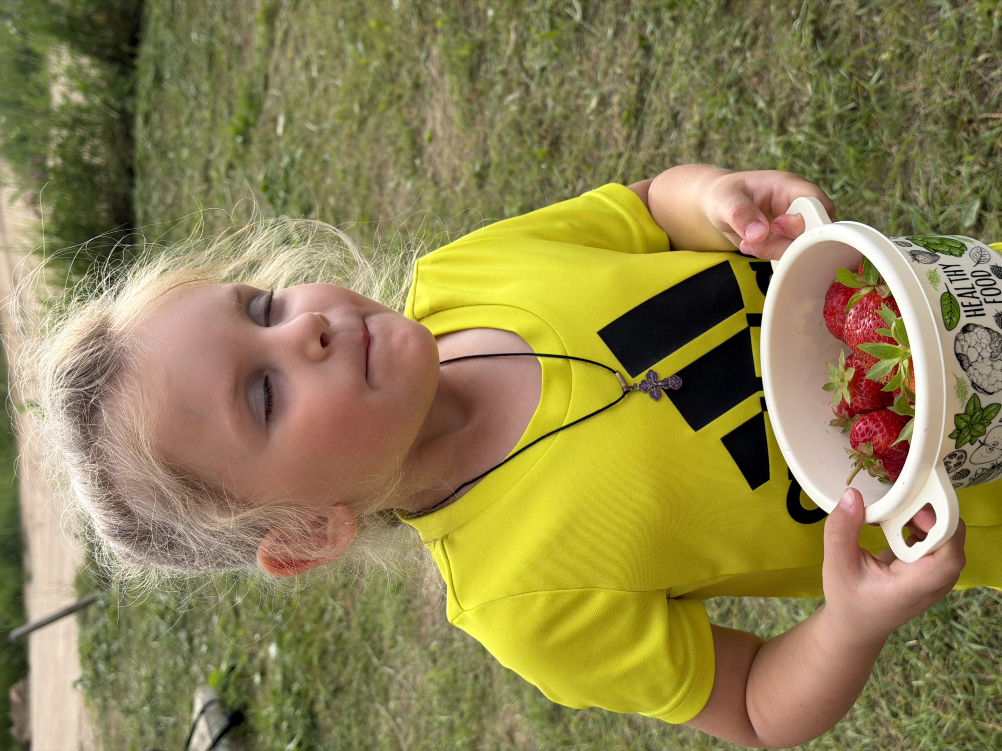 A young Caucasian girl with blonde hair, eyes closed, is holding a white bowl filled with strawberries. She is wearing a bright yellow t-shirt with a black Adidas logo and a dark necklace with a cross pendant. The girl appears to be outdoors in a grassy area. The background is blurred, showing more grass and some vegetation, suggesting a natural setting, possibly a park or a backyard. The image was taken in Chaikovsky, Russia.