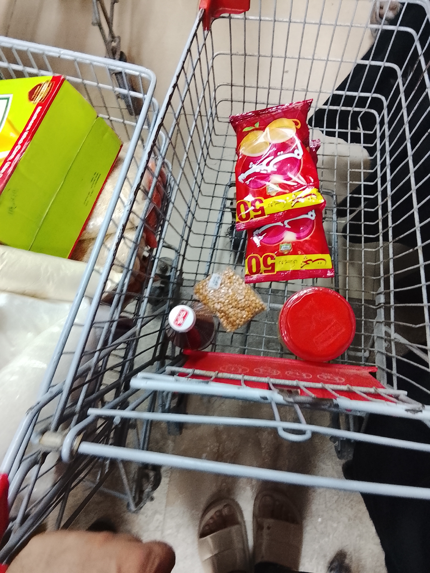 The image depicts a shopping cart in Gulgasht Colony, Pakistan, filled with groceries. A green and red box is in the first cart. In the main cart, there are two red bags of chips marked with the number "50" in Arabic, a small bag of peanuts, a bottle of liquid with a red and white cap, and a red round container with a lid. A person wearing beige sandals is standing near the cart. A person wearing a black abaya appears to be standing on the right side of the cart. Part of a person's hand is visible at the bottom of the image. The floor beneath the cart is light-colored.
